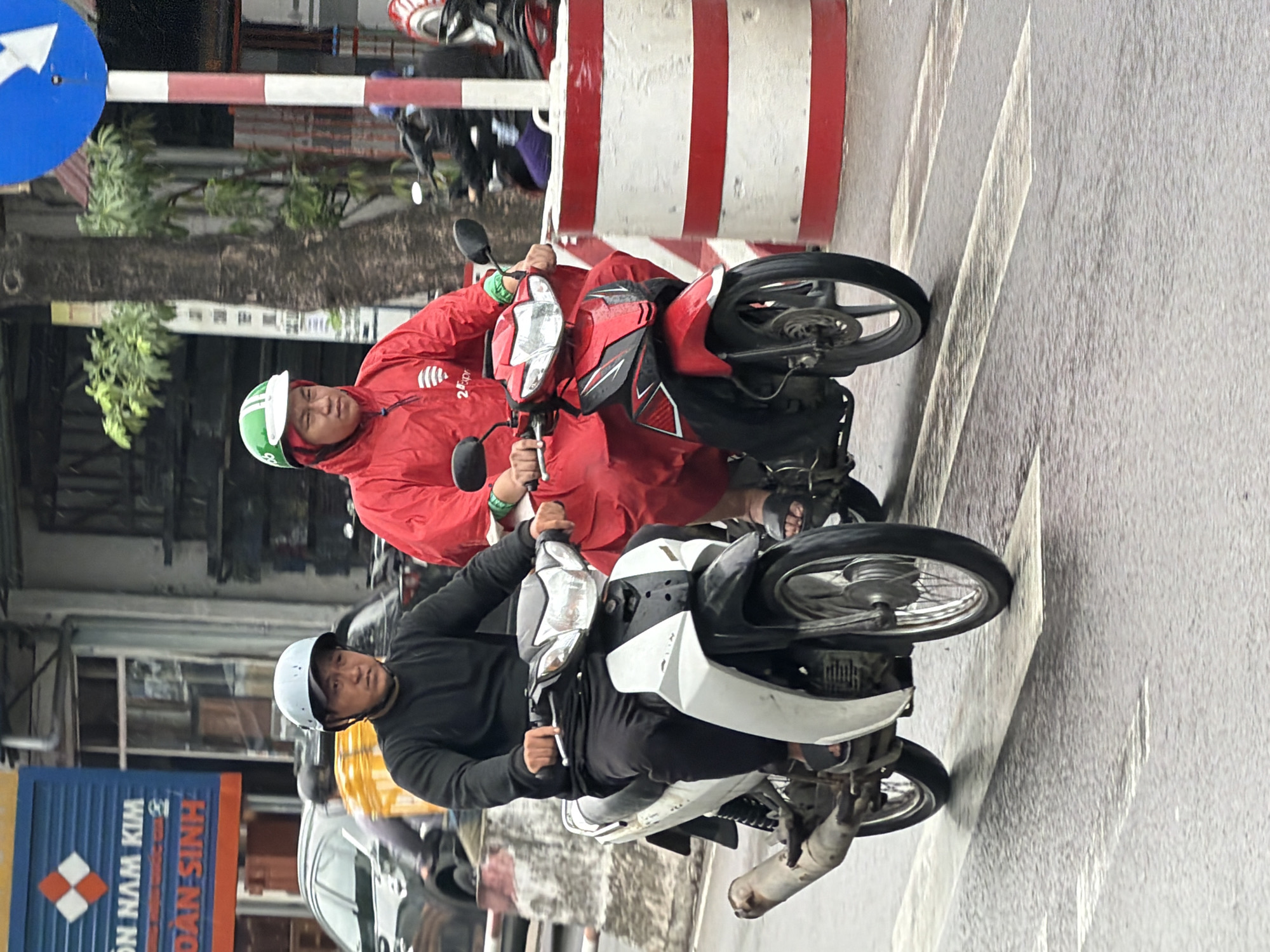 In Nha Trang, Vietnam, two individuals are riding motorcycles on a wet street, likely during daylight. The person in the foreground, wearing a light blue helmet and a dark long-sleeved shirt, is operating a white and black motorcycle. To their right, another person, distinguished by a green and white helmet and a bright red rain jacket with a logo, rides a red and black motorcycle. Both riders are wearing dark pants and sandals. The road surface appears damp, with white pedestrian crossing lines visible. In the background, a large red and white striped barrier stands near a tree. A blue street sign with white arrows is also present. Buildings and other vehicles are faintly visible further in the distance.