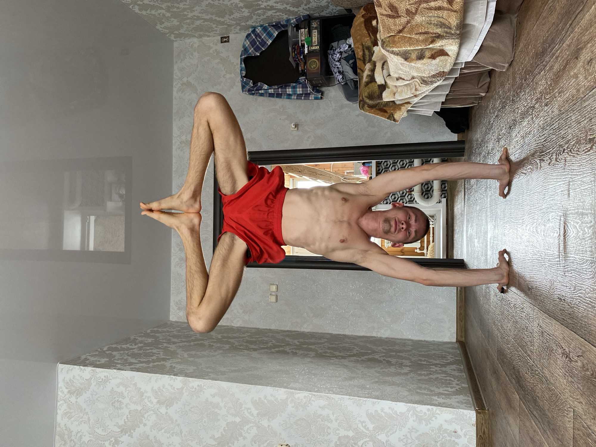 The main image, rotated 90 degrees counter-clockwise, captures a lean young man performing a handstand indoors. He is shirtless and wears red shorts, looking directly at the camera with a neutral to slightly strained expression. His hands are planted on a light-brown wooden laminate floor, which occupies the right side of the frame, while his legs are bent at the knees and extended upwards.
On the left side of the image, a light grey wall with a subtle damask-patterned wallpaper is visible. A prominent, highly reflective surface, likely a large mirror, runs along this wall, distinctly reflecting the sole of the man's left foot. This reflective surface extends towards the ceiling, which features the same patterned wallpaper. Two light-colored square electrical switches are mounted on the patterned wall.
In the background, above a dark-framed doorway, there is a cluttered storage area or wardrobe. A blue and white plaid shirt is draped over an item, and various objects, including books and a board game box clearly labeled "ROOT," are stacked. A brown, patterned blanket or throw is also visible. Through the doorway, some light from another room or outdoors is faintly discernible. The scene is brightly lit, indicative of daytime. This activity takes place in an indoor room located in Ulyanovsk, Russia.