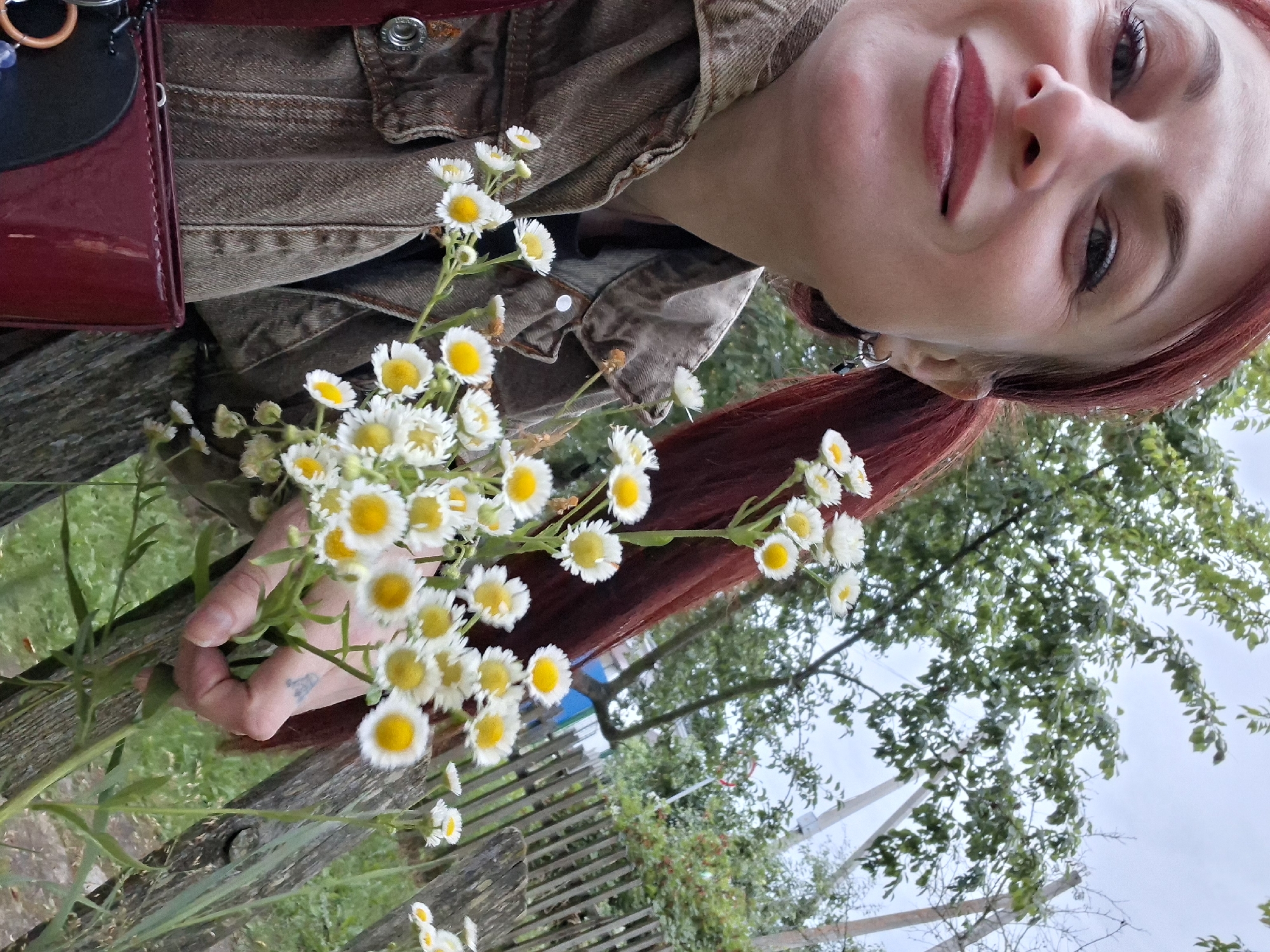 A woman with auburn hair tied in a ponytail is taking a selfie in an outdoor setting, possibly in Забалоцкі сельскі Савет, Belarus. She is wearing a denim jacket and holding a bouquet of small white and yellow flowers. The background features trees, a wooden fence, and foliage, suggesting a rural or garden environment. The woman is smiling and looking towards the camera.