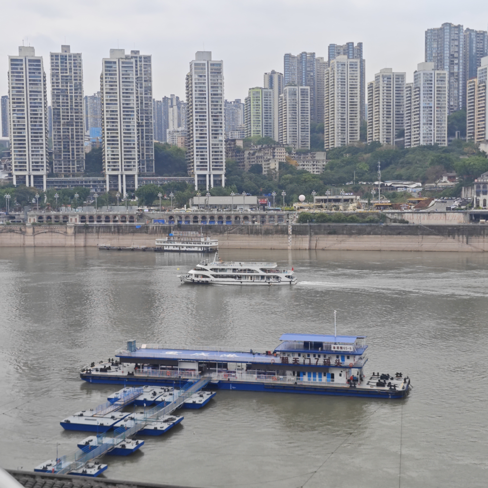 The image depicts a riverfront scene in Yuzhong District, China. In the foreground, a blue and white two-story ferry terminal platform is attached to multiple smaller blue and white floating platforms connected by pedestrian bridges. Midground contains two boats traveling in the water. The background features a cityscape with numerous high-rise apartment buildings and a waterfront infrastructure along the river. The weather appears overcast.