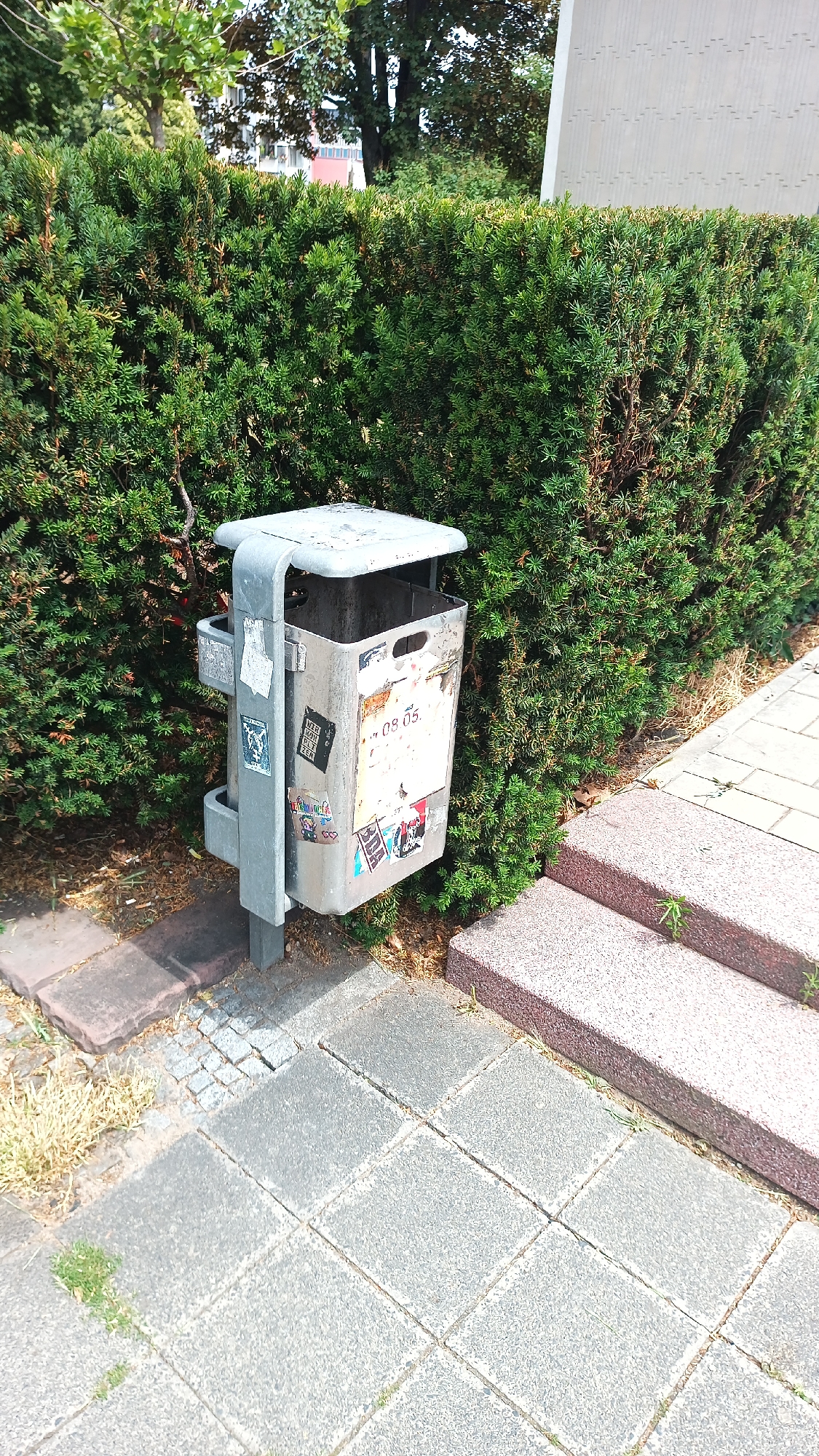A metal trash receptacle is positioned in front of a dense green hedge. The trash receptacle has a weathered appearance and is adorned with several stickers. A concrete sidewalk, composed of large square tiles, extends from the lower left to the lower right of the image. To the right of the sidewalk, there are two stone steps. In the background, trees with green foliage are visible, with a building and its facade showing. The scene is set on a sunny day, indicated by the lighting and shadows. The image was captured in Nuremberg, Germany, based on the available metadata.