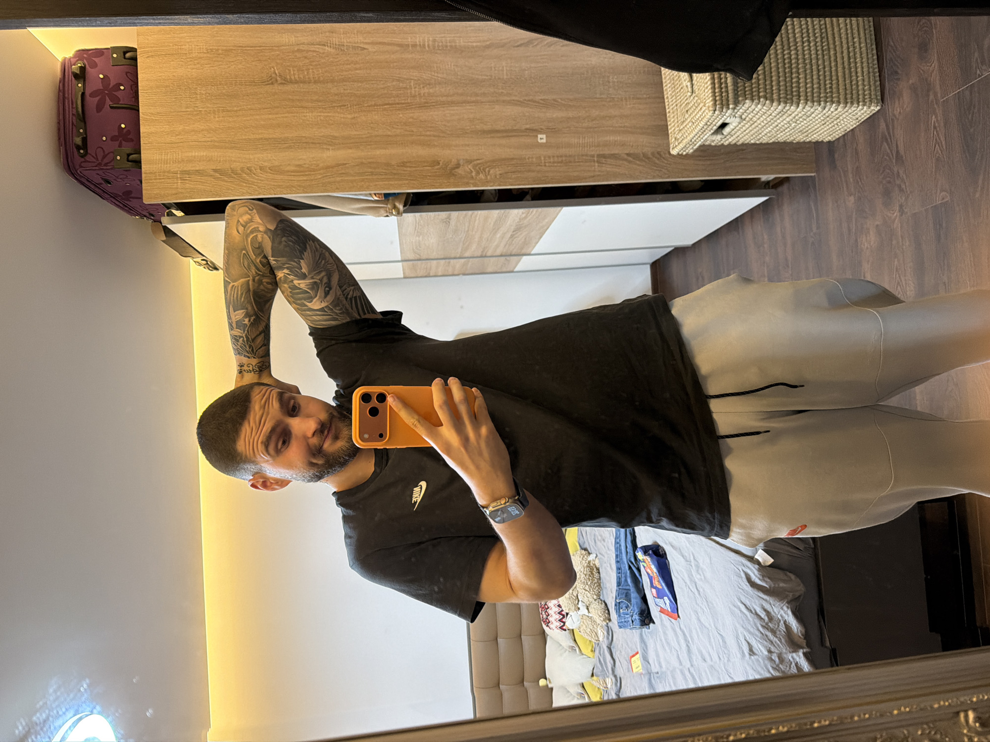 A man is taking a selfie in a mirror. He is wearing a black t-shirt and light-colored sweatpants. He has tattoos on his left arm. The background shows a room with a bed, nightstand, closet, and a suitcase. The location is in a residential area in the city of Sofia, Bulgaria.