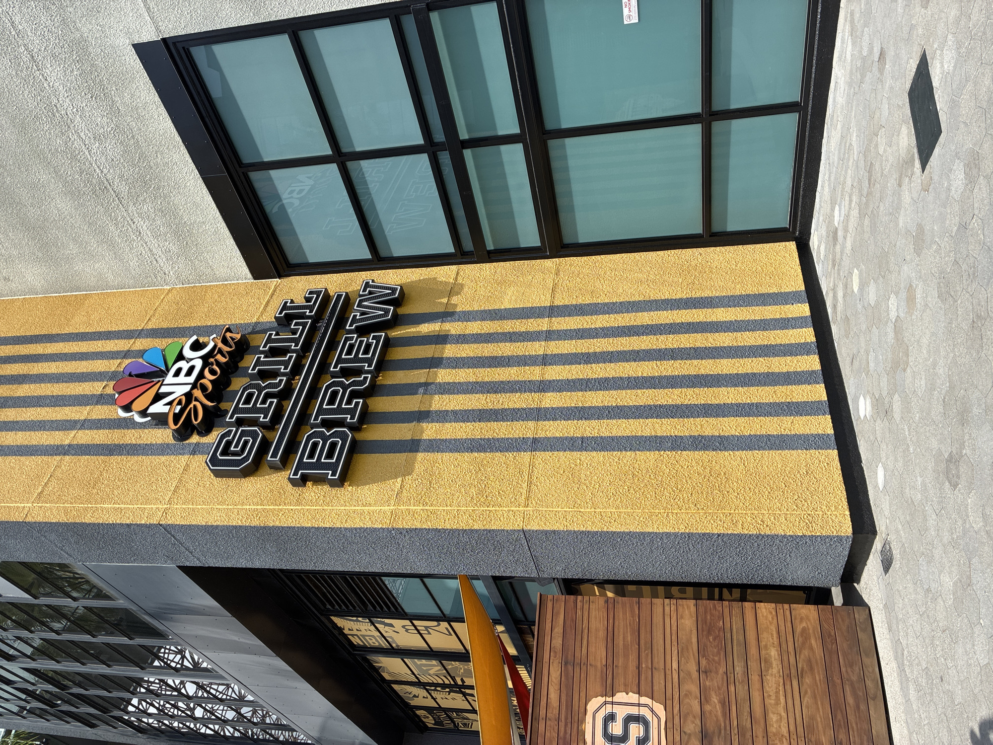 The image shows the exterior of an NBC Sports Grill & Brew restaurant. The building features a textured yellow wall with vertical gray stripes. The NBC Sports logo is prominently displayed in colorful, 3D lettering above the words "GRILL BREW" which are also in 3D black lettering with white outlines. To the left, a wooden structure with horizontal slats has a logo with the letter "S" inside a shield. To the right of the yellow wall, there is a large window with a black frame, divided into multiple panes, with a frosted or tinted appearance. The ground in front of the building is paved with hexagonal gray stones. The overall scene is an outdoor urban environment on a sunny day.
