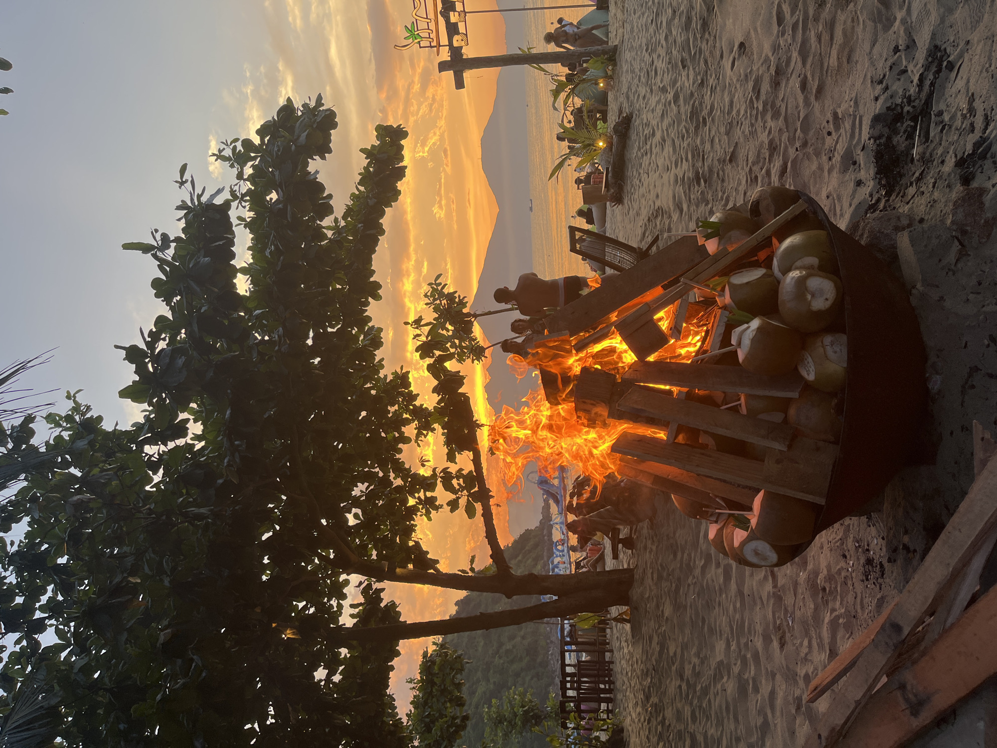 The image depicts a beach scene in Đà Nẵng, Vietnam, during sunset. In the foreground, a large bonfire constructed from wooden pallets is burning intensely, with fresh coconuts stacked around and within the structure. To the left of the fire, a tree with dense foliage partially obscures the view.
In the background, the sunset casts a warm, orange glow across the sky and the mountainous terrain. Several people are visible in the background; some appear to be seated at tables or lounging on the beach, and others are standing near a beachside structure with a decorative sign. The scene is set on a sandy beach with a slight slope towards the ocean, which is dimly visible in the distance.