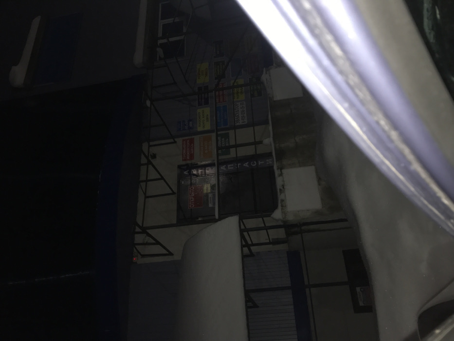 Captured from a skewed perspective, the image reveals a building façade at night. The building exhibits a metal frame adorned with various advertisements and notices. A storefront with signage in a Cyrillic script is visible. The scene is dimly lit, with the darkness amplifying the visual impact of the lit advertisements. Part of a car appears in the foreground.