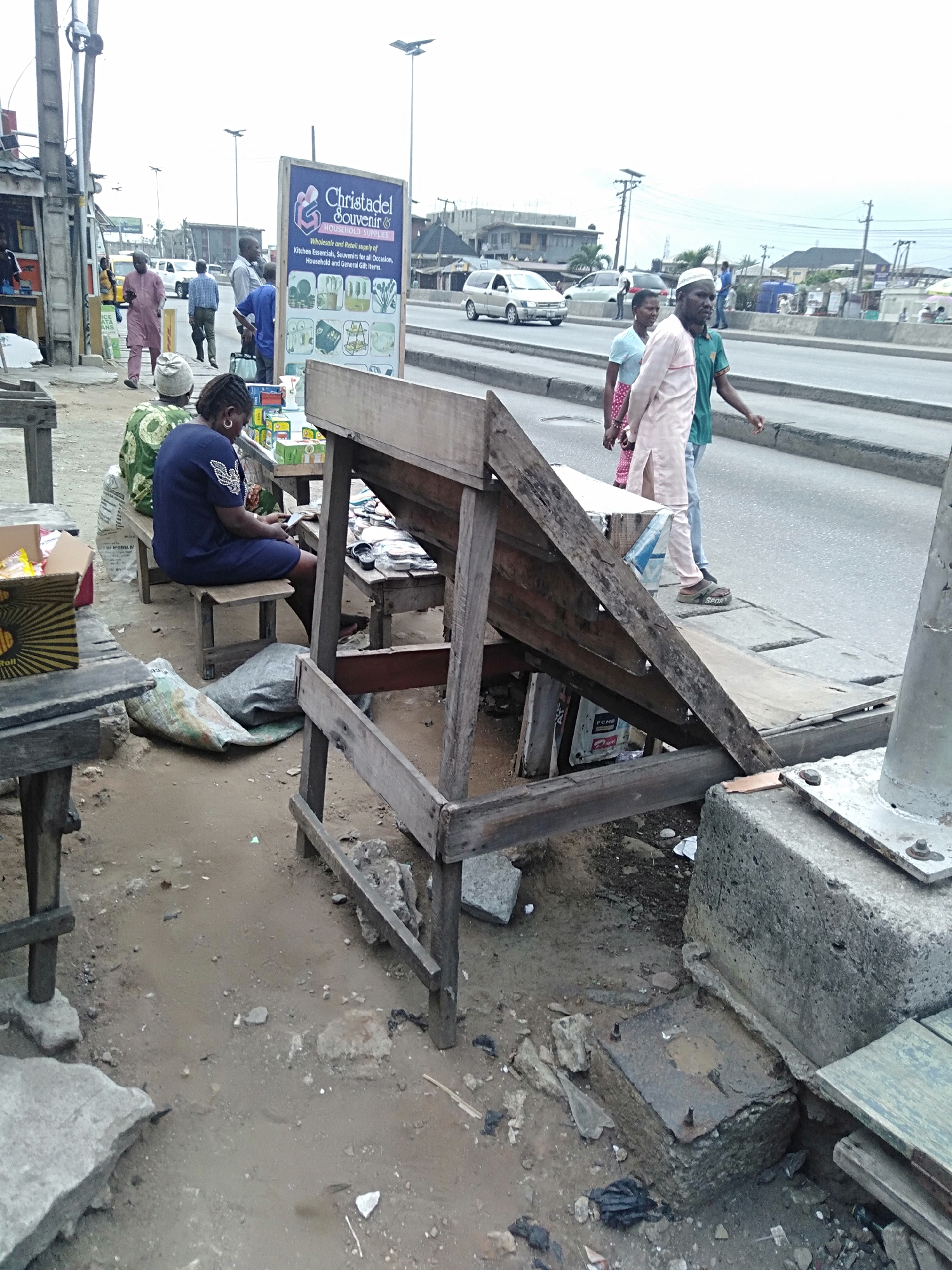 A daytime shot taken at street level in Shangisha, Nigeria, features a busy street scene. A main road runs horizontally through the background, with several vehicles and pedestrians visible. On the left side of the image, there's an open-air market setting. People are seated on benches, some appearing to be engaged with their mobile phones. There are makeshift wooden stands and displays of goods, though the specific items are not clearly discernible. The foreground is dominated by weathered wooden structures, possibly part of market stalls. Debris litters the ground. A sign for "Christadel Souvenir HOUSEHOLD SUPPLIES" is visible in the background. Several individuals are present, wearing varied clothing styles. A man in a pink robe and a white cap is among them, walking alongside others near the main road. The sky overhead is overcast.