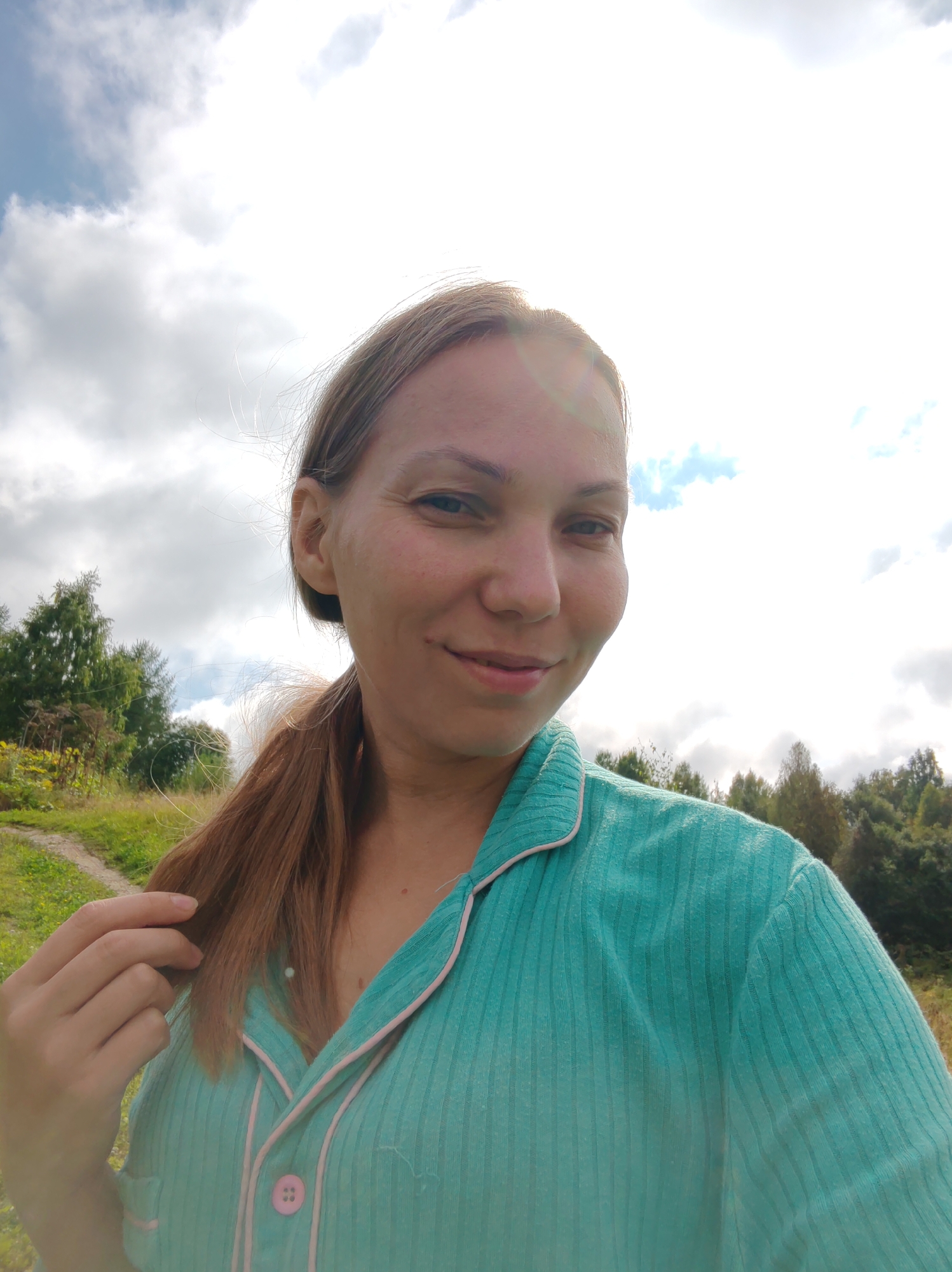 The image shows a woman taking a selfie outdoors. She is looking towards the camera with a slight smile. Her hair is pulled back into a ponytail, and she is wearing a teal colored shirt with pink trim and a pink button. The background consists of a cloudy sky, trees, and a grassy path. The location is described as the city of Чухлэм, Russia.