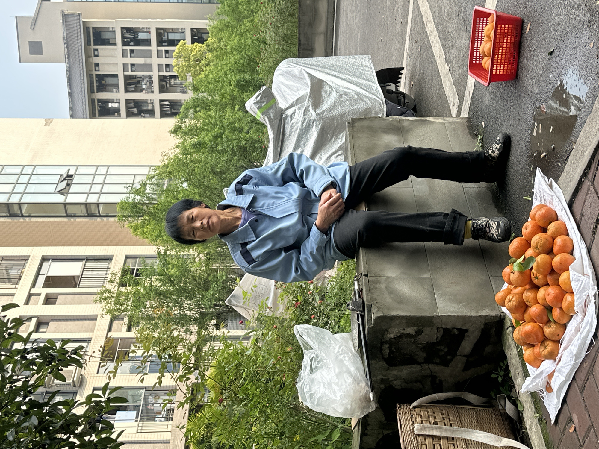 A horizontal, eye-level shot shows an Asian woman sitting on a concrete structure in Jiangjin, China. She wears a light blue jacket, dark pants, and patterned sneakers, looking at the camera with a slight smile. Her hands are clasped in her lap.
Beside her are piles of oranges in white bags, and a red basket containing oranges sits on the wet pavement. A covered motorcycle is visible behind her, and a building with many windows stands in the background along with green trees. A wicker basket and a plastic bag are on the ground beside the concrete structure.