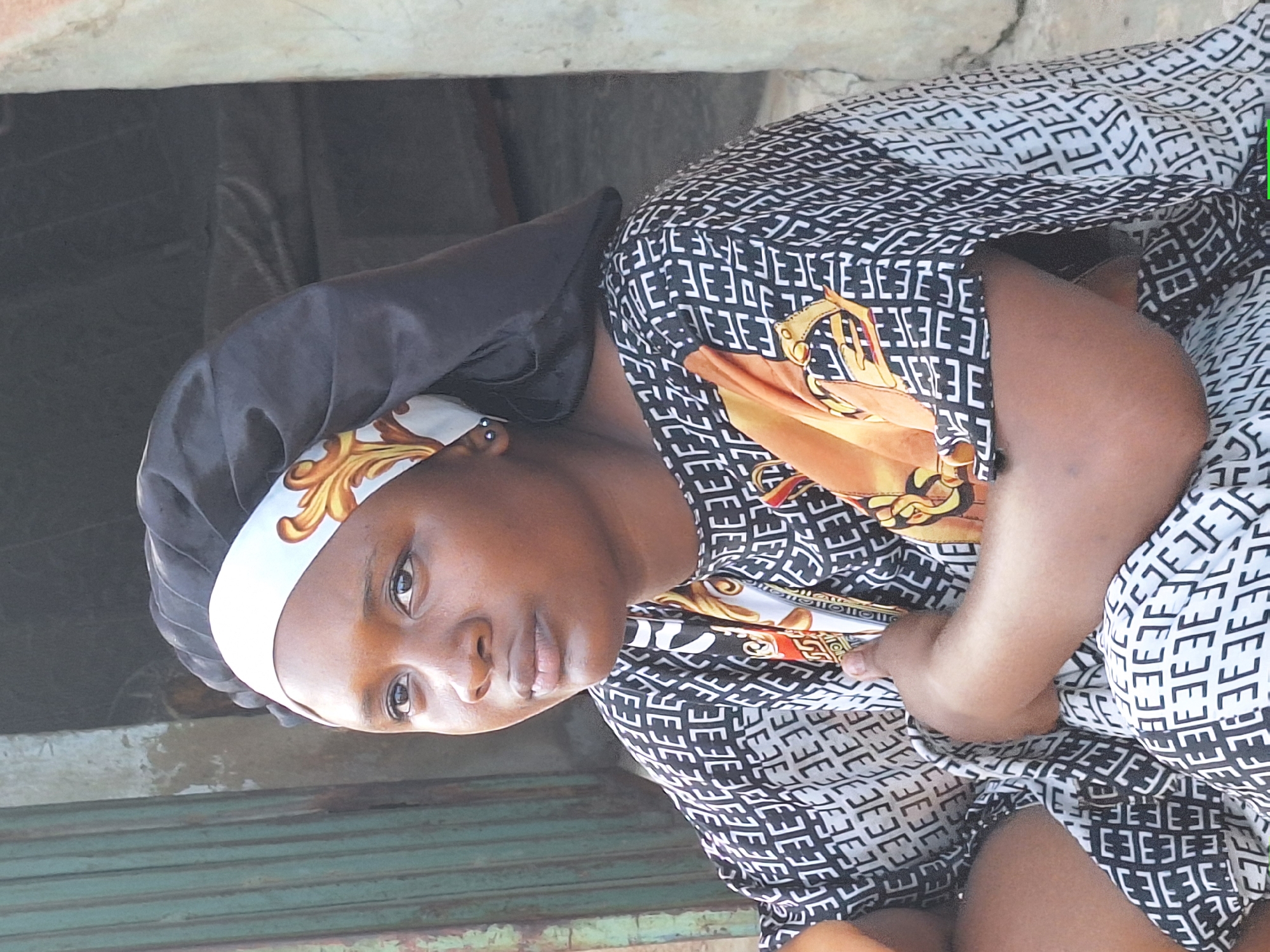 A woman sits indoors in Sabon Tasha, Nigeria. She wears a black headscarf with a white and gold patterned band and a patterned black and white dress with gold detailing. She is looking directly at the camera. The background includes a concrete wall and what appears to be a green roll-up door.