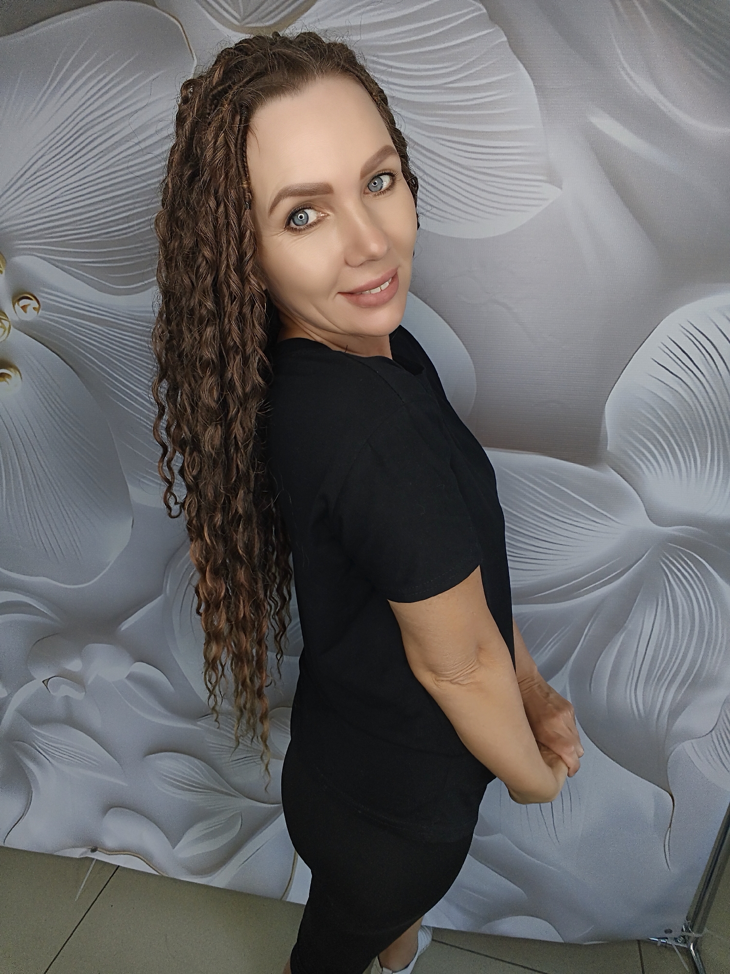 In an indoor setting with a large, white floral-patterned backdrop, a woman is captured in a three-quarter profile view, looking towards the camera. She has long, dark, curly hair styled in braids, and striking blue eyes. She is wearing a black t-shirt and black pants. Her arms are clasped in front of her. The lighting appears to be natural or soft artificial light, illuminating the subject and casting subtle shadows. The overall scene is neutral and focused on the woman.