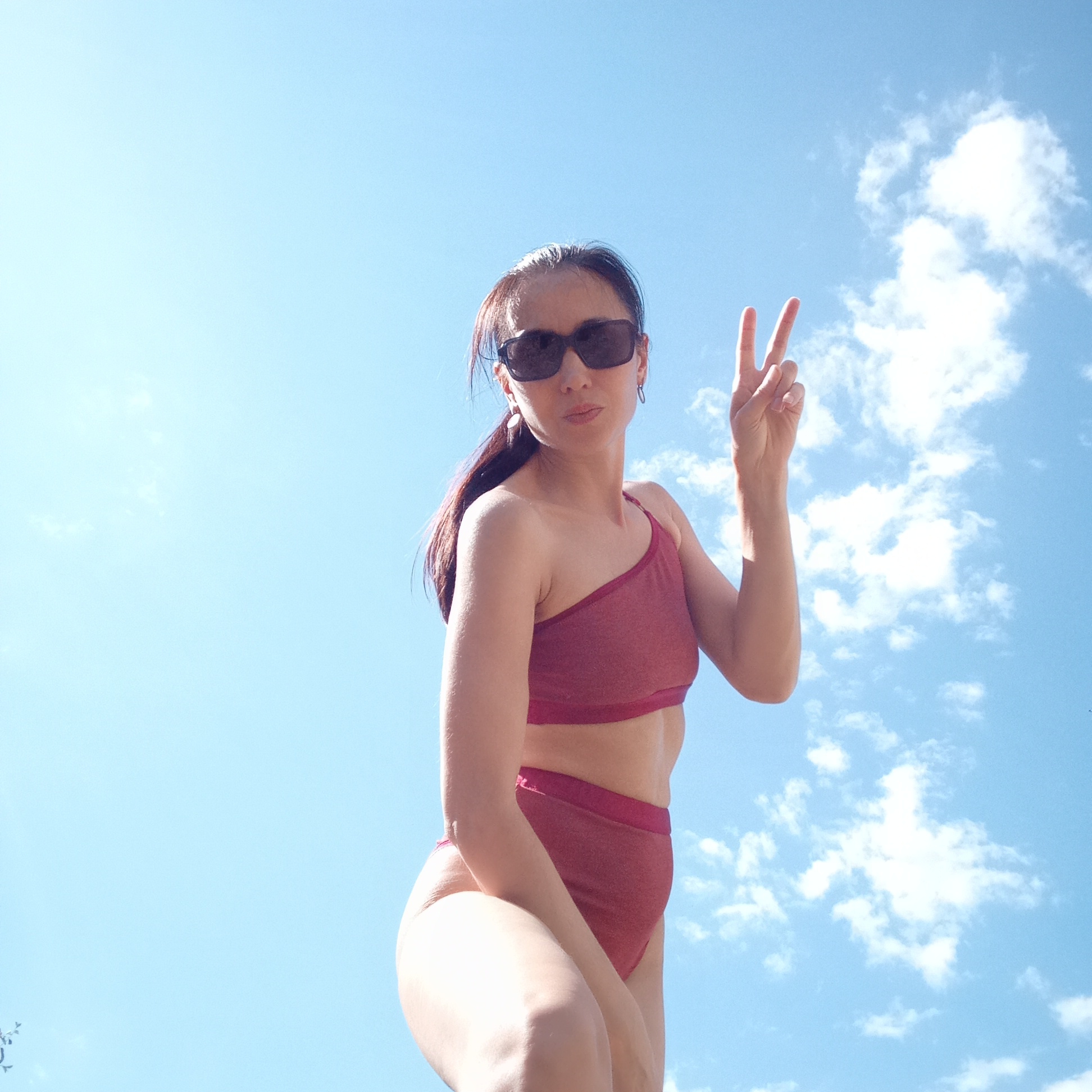 A woman with sunglasses is standing outside under a blue sky with scattered clouds, making a peace sign with her right hand. She is wearing a maroon one-shoulder swimsuit. The location is unspecified and there is no mention of the city of Буренка, Russia in the image.