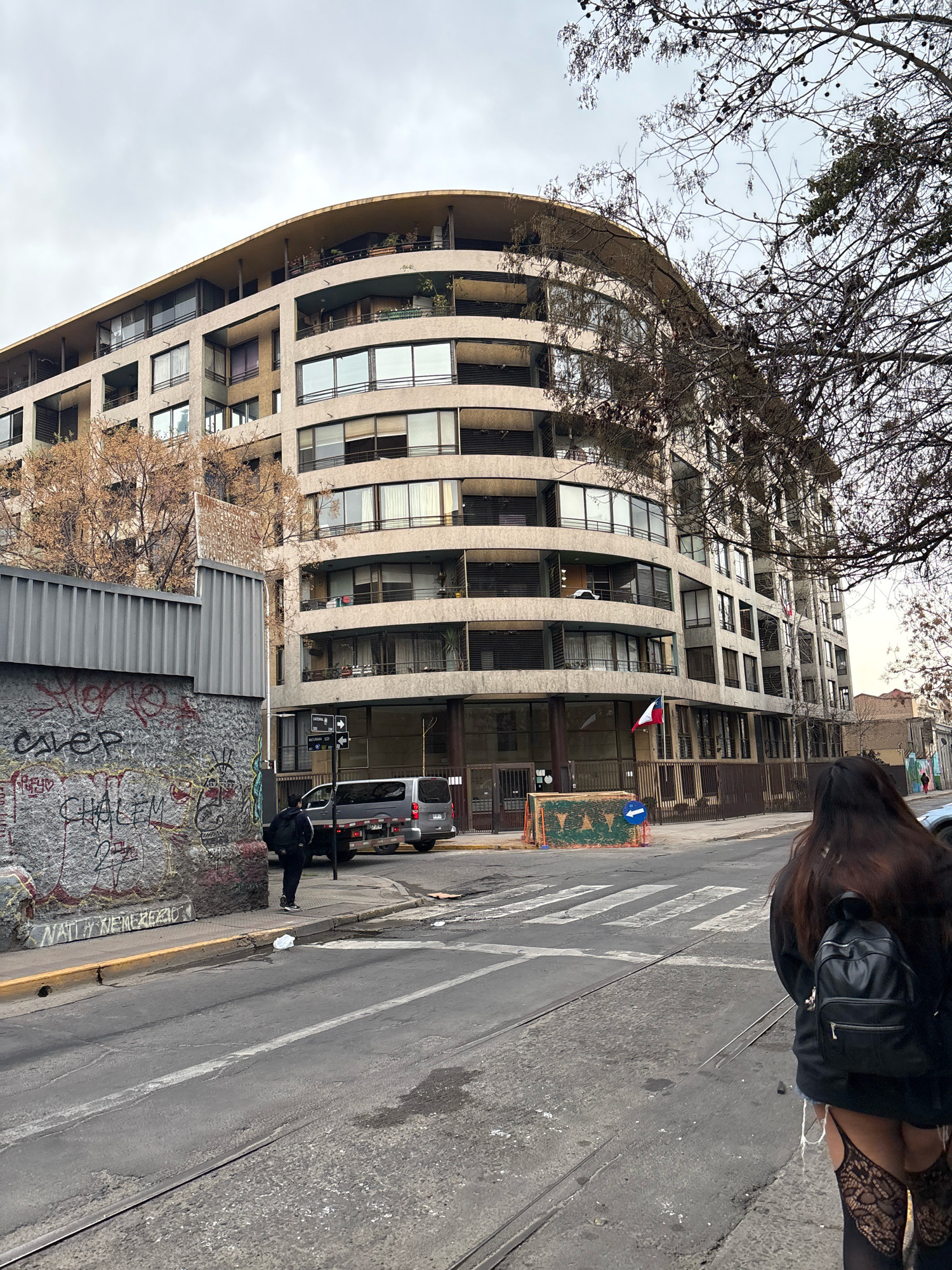 The image depicts a street scene in Santiago, Chile, with a modern, curved apartment building dominating the background. The building, made of concrete and glass, has numerous balconies with railings and large windows. A flag of Chile is visible on the building. In the foreground, a street with tram tracks runs horizontally across the image. A gray van is parked on the right side of the street. A stone wall covered in graffiti is on the left side of the street. A person wearing a black jacket and backpack, and lace stockings, is walking away from the camera on the right sidewalk. The sky is overcast.