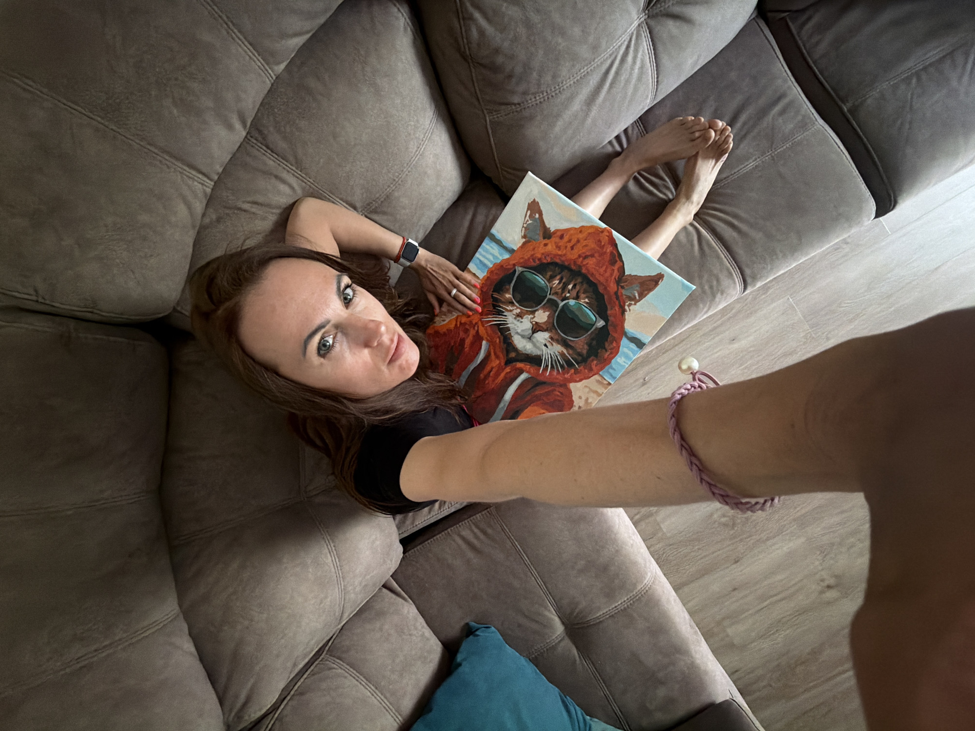 A woman is depicted lying on a large, grey-brown sectional sofa, capturing a selfie from an overhead perspective. Her head is positioned towards the upper left, with her body extending across the sofa. She has long, dark brown hair and is wearing a black top. Her eyes are light-colored and directed towards the camera.
On her left wrist, she wears a red-strapped smartwatch, and a ring is visible on her finger. She is holding a rectangular painting on her chest. The painting features a stylized cat wearing a red hooded sweatshirt and reflective round sunglasses, set against a background depicting a light blue sky and a sandy beach. Her bare legs and feet are visible to the right of the painting, resting on the couch.
Her right arm is extended, presumably holding the camera, and a braided light pink bracelet with a white pearl-like accent is visible on her wrist. The sofa has distinct stitched patterns on its cushions. A wooden floor is visible in the background, and a teal-colored decorative pillow rests on the couch near her extended arm. The scene portrays an indoor setting in Birobidzhan, Russia. No visible text is present in the image.
