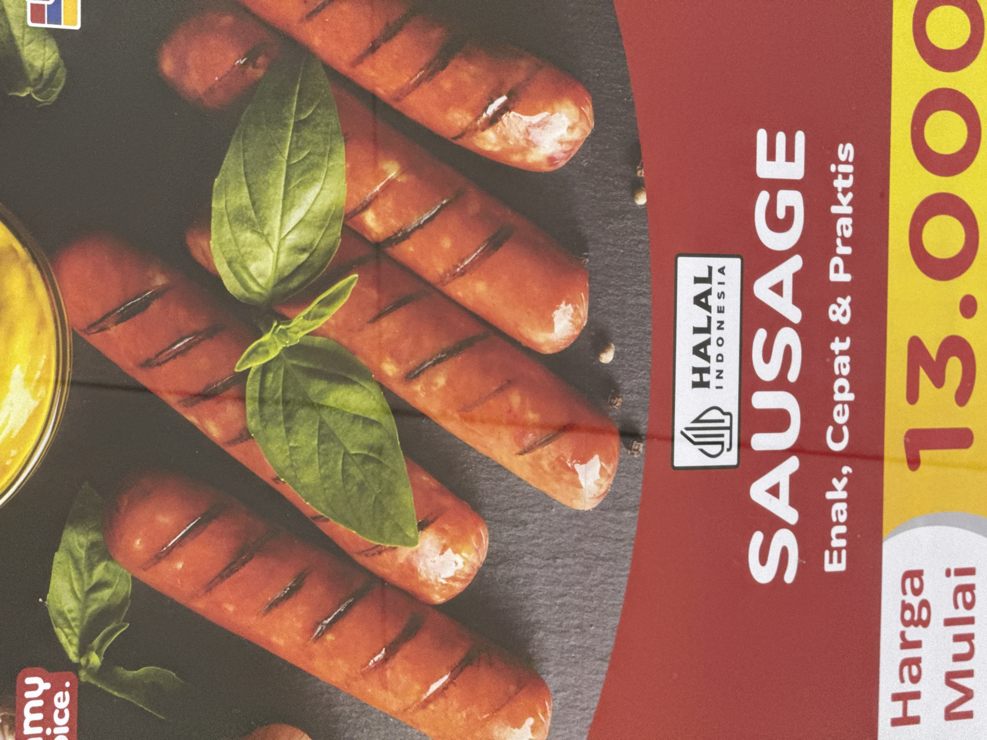 The image shows a package of sausage with the words "SAUSAGE," "Enak, Cepat & Praktis," "Harga Mulai," and "13.000" printed on it. The sausage is topped with basil leaves. The package also has a "HALAL Indonesia" logo. It is not possible to determine the location as Kuta, Indonesia, from the image.
