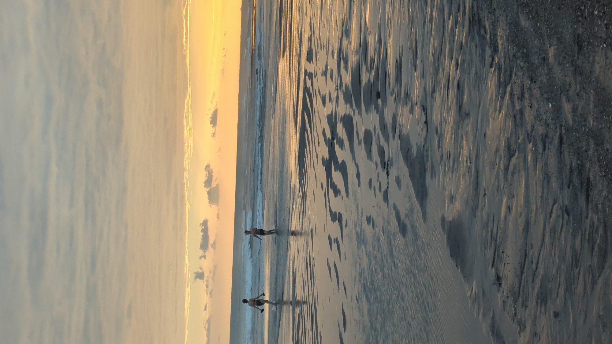 The media file depicts a serene beach scene at sunset in North Kuta, Indonesia. Two individuals are walking along the wet sand, their reflections stretching out before them. The setting sun casts a warm, golden glow across the sky, illuminating the scattered clouds and creating a tranquil atmosphere. The gentle waves of the ocean lap at the shore, their movement captured in the rippling patterns on the sand. The sand itself is a mix of light and dark tones, with intriguing textures formed by the receding tide. The overall mood is one of peace and quiet contemplation, inviting viewers to imagine themselves in this idyllic location. There is no visible text in the media file.