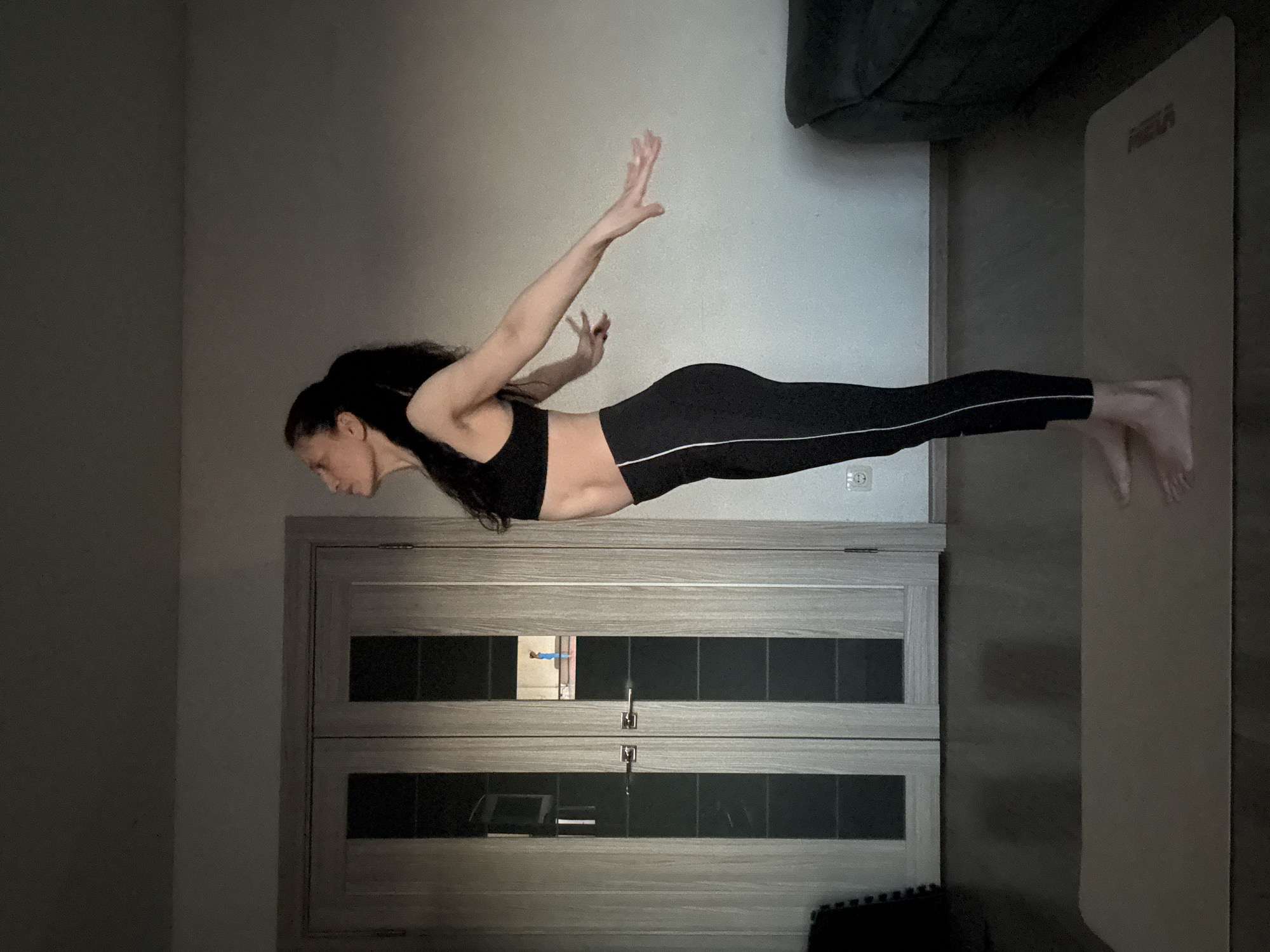 A woman is captured mid-exercise indoors, performing a challenging yoga or Pilates pose, resembling a "superman" or "locust" variation. She is suspended horizontally, parallel to the ground, with her body perfectly straight from head to toe. Her arms are extended backward and slightly upward, hands open, while her legs are held together and extended straight. She is dressed in black athletic wear, including a sports bra and form-fitting leggings with white stripes along the outer seams. Her dark, long hair is pulled back, and her facial expression shows concentration and effort.
The scene appears to be a room within a home, with soft, ambient indoor lighting suggesting it could be evening or a dimly lit space. The background features a plain light grey wall and a double door with grey wooden frames and dark glass panels, through which a faint reflection of a person in a blue top is visible. A white wall socket is also visible on the wall. To the right of the woman, a light-colored yoga mat is partially visible, with the word "MEGA" printed on it. A dark, possibly upholstered piece of furniture or a couch is partially seen in the upper right and left corners of the image. The entire image is rotated 90 degrees clockwise, making the woman appear horizontal, though she would be upright relative to the floor in the actual space. The location context is Chaikovsky, Russia.