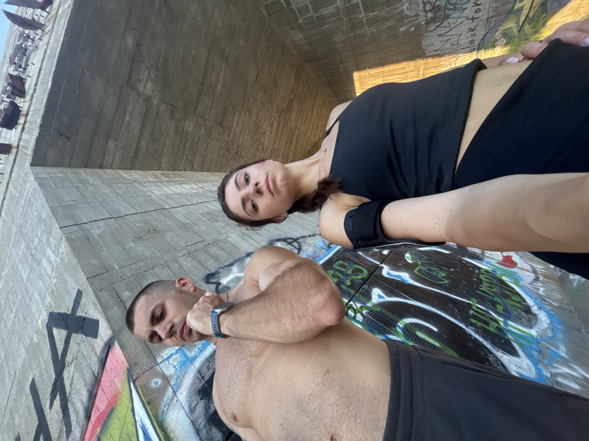 This media file features two individuals, a man and a woman, outdoors. The man, shirtless and wearing dark shorts and a watch, is positioned in the foreground, leaning against a concrete structure covered in graffiti. He has short hair and a muscular build. The woman, wearing a black tank top and dark athletic wear, is behind him, appearing to be lying down or reclining. She has dark hair tied in a ponytail.
The setting appears to be under a large concrete structure, possibly a bridge or overpass, with graffiti art adorning the walls and ground. The concrete is weathered and shows signs of age. The lighting suggests it is daytime, with sunlight casting shadows. The overall mood seems relaxed, possibly after a workout or during a break.
There is visible text in the form of graffiti on the concrete. Some legible phrases include "for" and "black". The location context provided is Varna, Bulgaria. The image is shot from a low angle, looking upwards towards the woman and then down towards the man and the graffiti-covered ground.