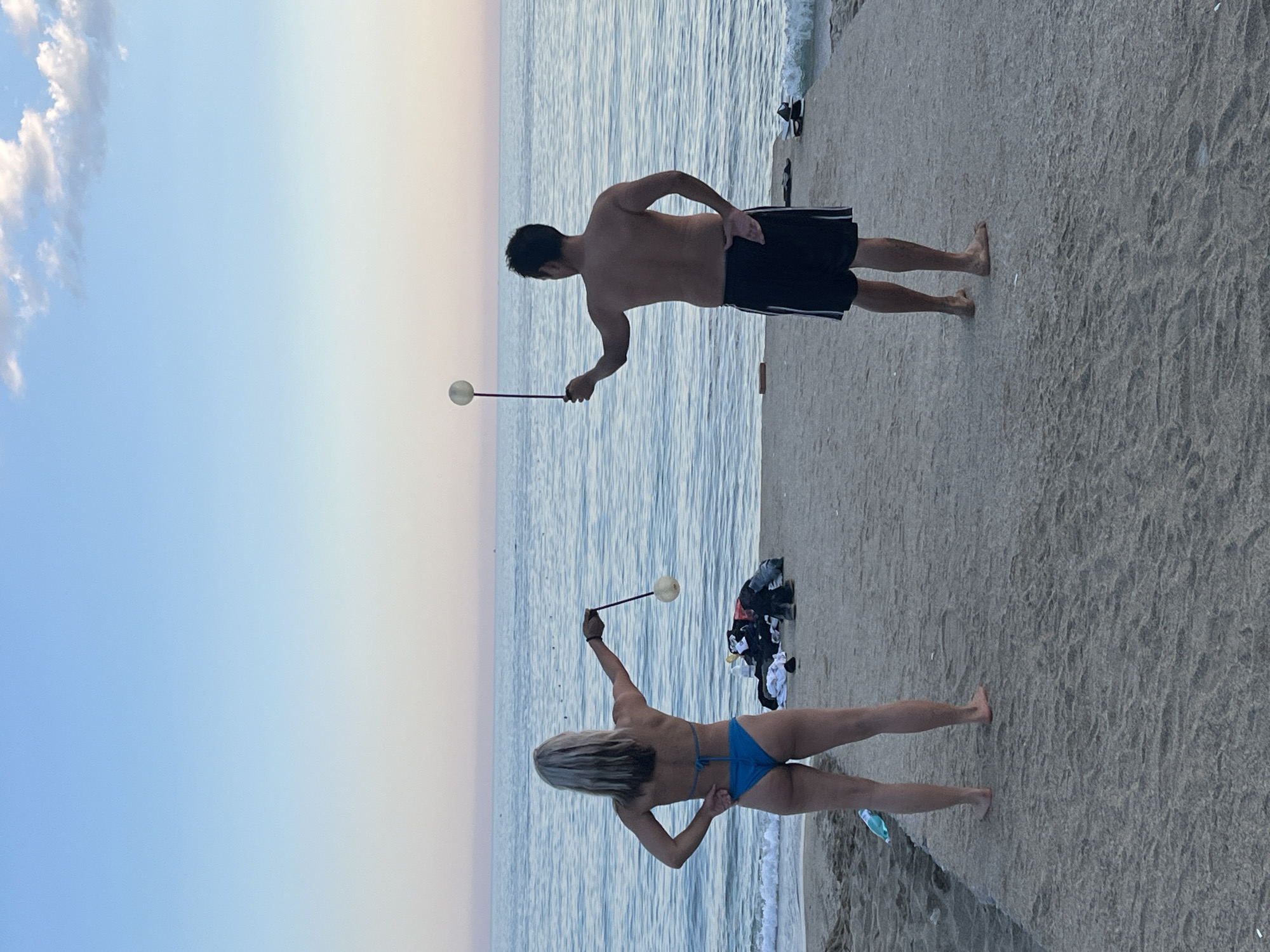 Two people, a man and a woman, stand on a sandy beach in Alanya, Turkey, illuminated by a setting sun. The man, shirtless and wearing dark shorts with white stripes, holds a white ball attached to a stick. The woman, wearing a blue bikini, mirrors his action with a similar ball and stick. Clothing is strewn nearby, and the sea gently laps against the shore. The sky transitions from a clear blue to a soft orange near the horizon.