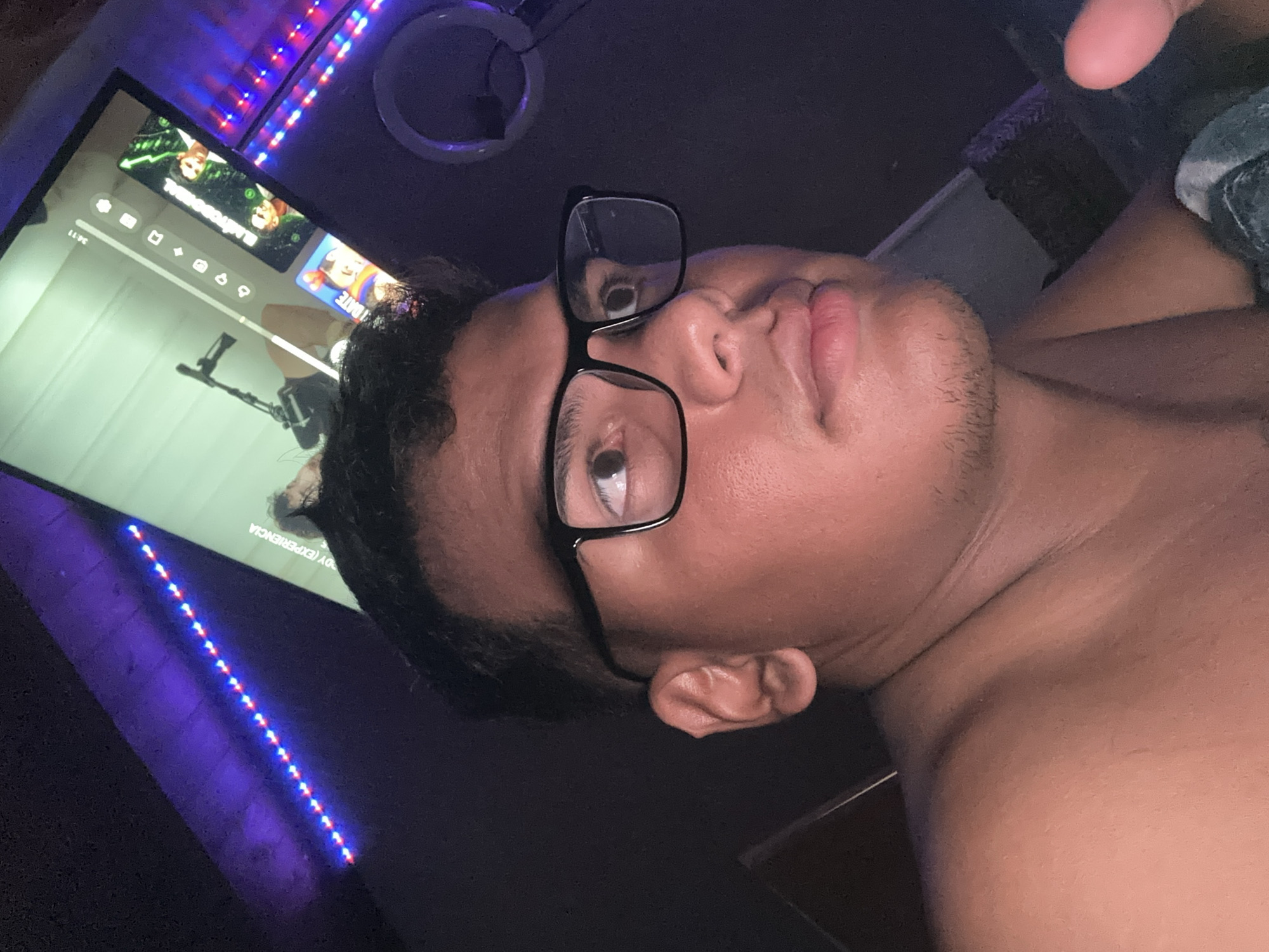 A close-up, angled photograph depicts a person with dark hair, wearing black-framed glasses, gazing toward the camera. The person's skin appears tan. The individual is partially unclothed, with a shoulder and part of the chest visible. Behind the person is a television screen displaying a variety of icons and images. A portion of a microphone stand is also visible on the screen. LED strip lighting, alternating red and blue, illuminates the area around the screen. A circular object hangs above the television. The location is not discernible from the image. There is no indication of a city, country, or location of any kind.