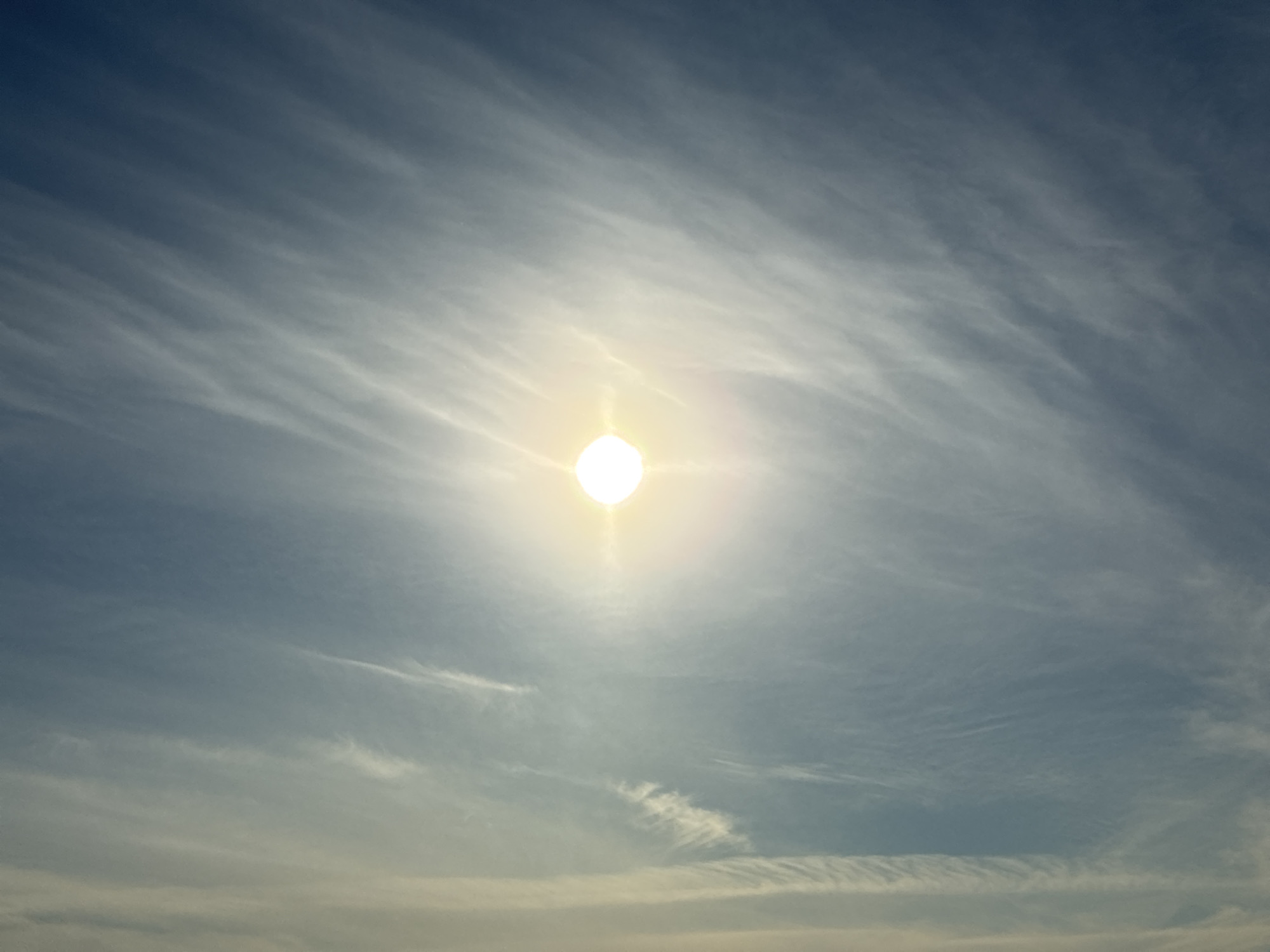 An extreme wide shot captures a bright sun in the center of a vast, clear blue sky. Thin, wispy clouds streak across the sky horizontally, creating a textured appearance. The sun is a bright, white orb with a faint halo. The bottom of the image shows a sliver of what appears to be land or a very distant horizon, blurred and out of focus.