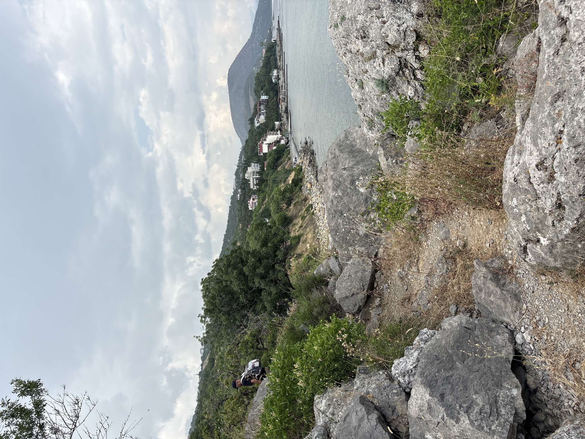 A man with a backpack is hiking on a rocky trail overlooking a coastline. The trail is lined with large gray boulders and dry grass. In the background, a body of water stretches to the horizon, and a town with white buildings is nestled along the coast. Lush green trees cover the hills behind the town. The sky is overcast with gray clouds. The location is Bondarenkovo, Ukraine.