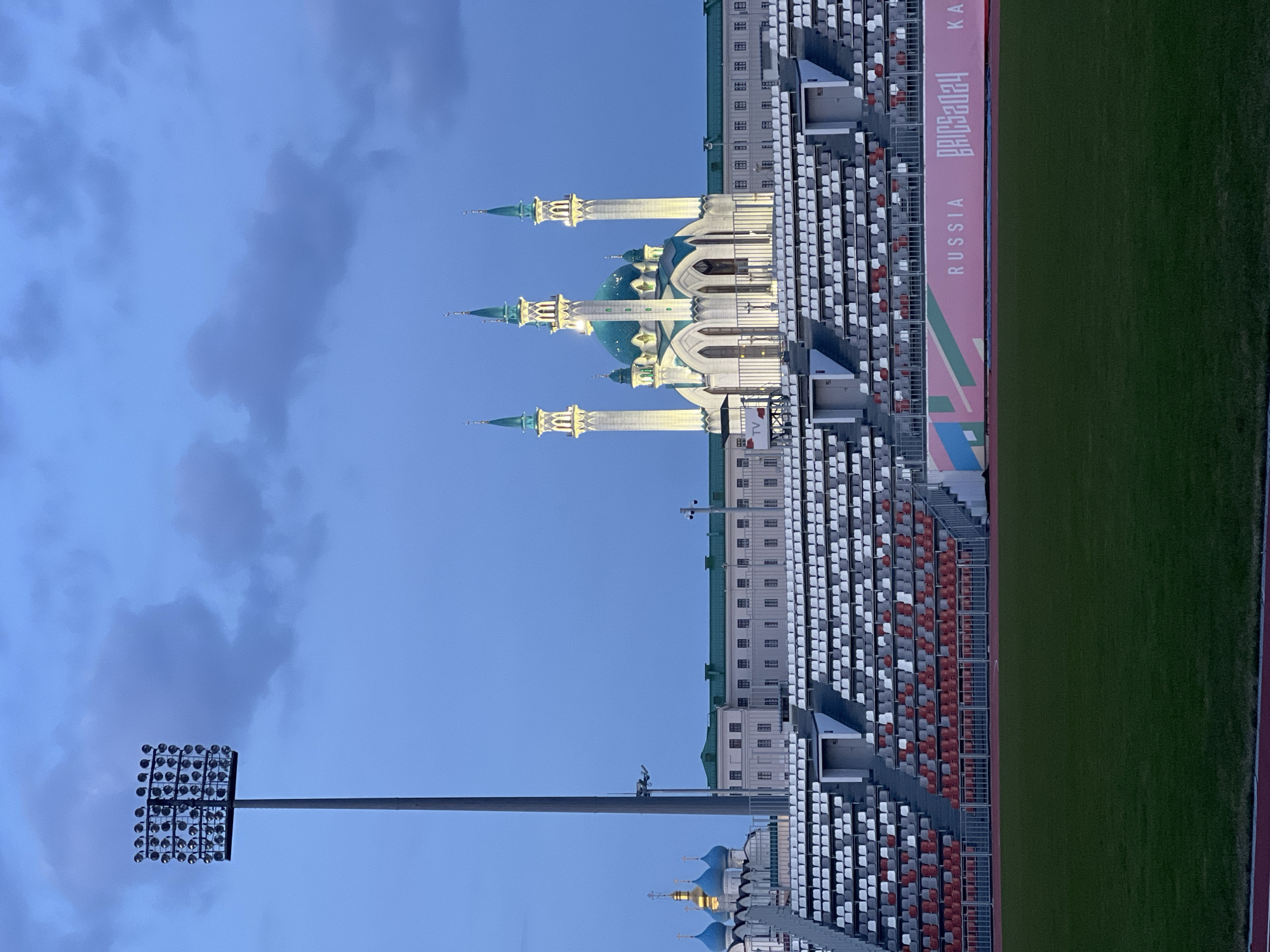 The image depicts a view in Kazan, Russia, featuring the Kul Sharif Mosque and the Kazan Arena. The mosque is distinguished by its four minarets and blue domes. Next to the mosque stands Kazan Arena, a soccer stadium with visible seating tiers and a green playing field. A large light array pole is to the left of the mosque. The stadium displays the words "RUSSIA BRICS 2024 KAZAN" on a banner. The sky is blue with cloud cover.