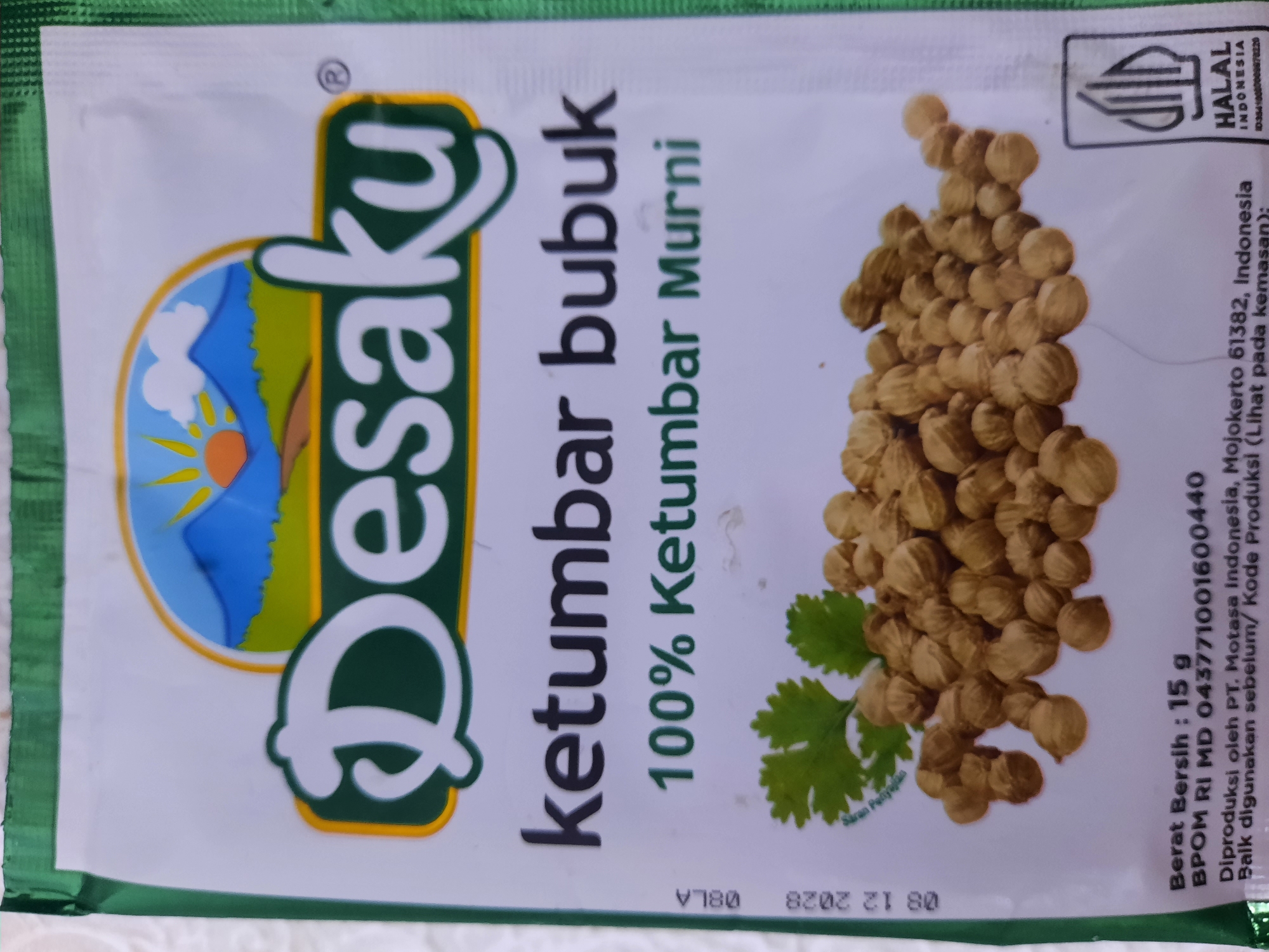 This is a close-up, high-angle photograph of the packaging for "Desaku Ketumbar Bubuk," a ground coriander spice product from Indonesia. The packaging is primarily white with green and yellow accents. A prominent green and yellow graphic features a stylized mountain landscape with a sun and clouds. To the right of this graphic, the brand name "Desaku" is written in large, bold, white letters with a green outline. Below the brand name, "ketumbar bubuk" (ground coriander) is written in smaller, black, vertical text, followed by "100% Ketumbar Murni" (100% Pure Coriander) in dark green, vertical text.
On the right side of the packaging, a cluster of whole coriander seeds is depicted, along with two green cilantro leaves. A small, stylized green leaf graphic is visible near the bottom right corner of the coriander seeds, with faint text above it that appears to say "Saran Penyajian" (Serving Suggestion).
In the upper right corner, there is a Halal certification logo for Indonesia, with text that reads "HALAL INDONESIA" and an identification number.
Below the image of the coriander seeds, there is text providing product details. It states "Berat Bersih: 15 g" (Net Weight: 15 g), followed by the BPOM (Indonesian National Agency of Drug and Food Control) registration number, "BPOM RI MD 043771001600440". Further down, it indicates the producer: "Diproduksi oleh PT. Motasa Indonesia, Mojokerto 61382, Indonesia" (Produced by PT. Motasa Indonesia, Mojokerto 61382, Indonesia). The text also includes instructions regarding shelf life and production code: "Baik digunakan sebelum/ Kode Produksi (Lihat pada kemasan)" (Best used before/ Production Code (See on packaging)).
At the very bottom of the packaging, a date stamp is visible, "08 12 2028", and a code "V780". The overall presentation suggests a product intended for culinary use, highlighting its purity and origin. The photo was likely taken in Lhokseumawe, Indonesia, as indicated by the location context. There are no people or specific environmental details like time of day or weather visible in the image.