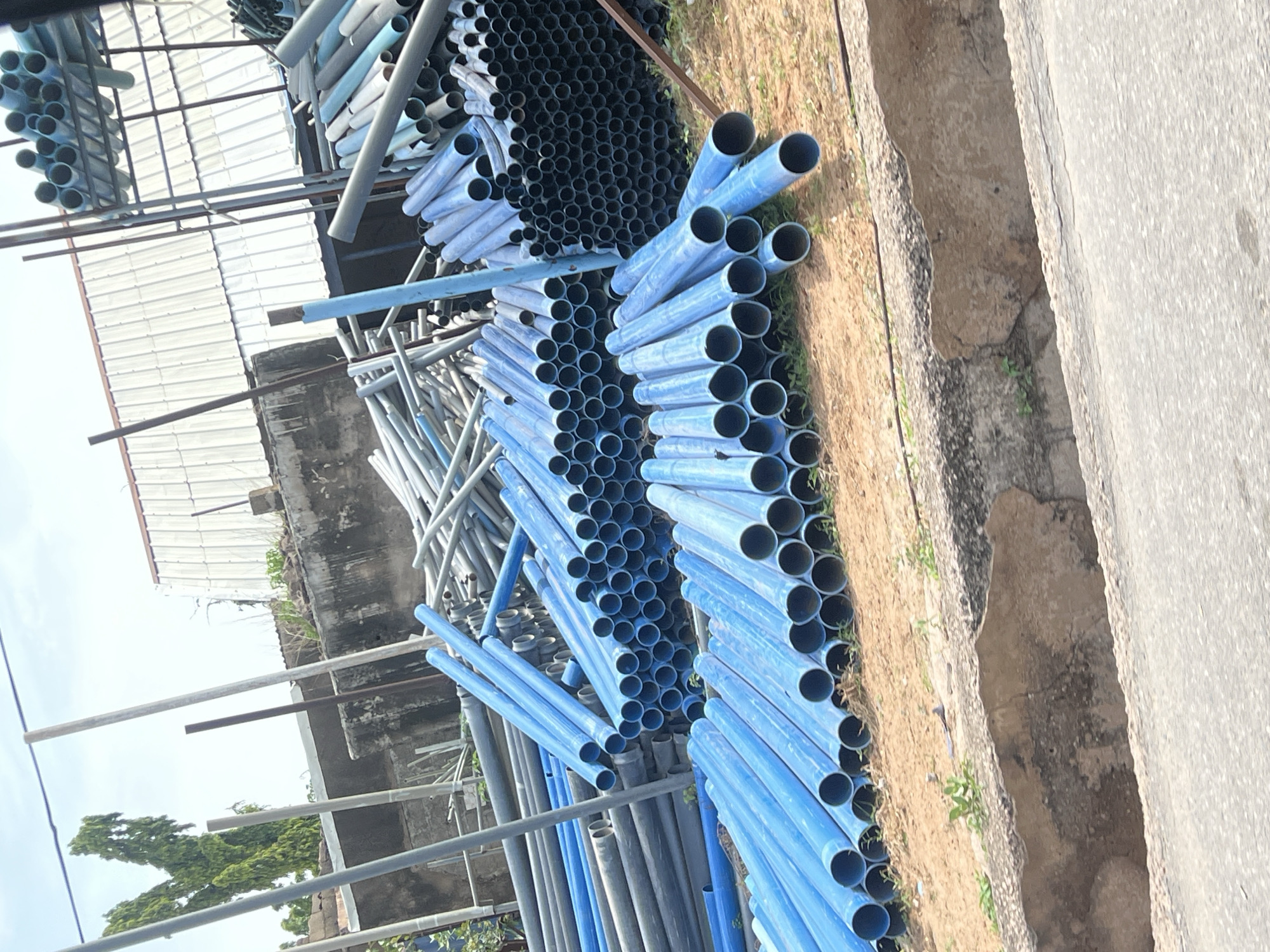 This media file displays a collection of blue and grey pipes, likely made of PVC or metal, stacked outdoors. The pipes vary in size and diameter, with some lying horizontally and others stacked vertically. They appear to be stored in a yard or construction site, with visible elements of a building structure and a dirt ground on one side, and a paved surface on the other.
The scene is illuminated by natural light, suggesting it is daytime. The lighting and shadows do not indicate a specific time of day like dawn or dusk. There are no people visible in the image, and the focus is entirely on the pipes and their surroundings. The overall impression is one of an industrial or construction-related setting. The context provided, "Jalingo, Nigeria," places this scene geographically. There is no visible text in the image. The arrangement of the pipes suggests they are either for sale, awaiting installation, or are surplus materials. The pipes are the primary objects of interest. The blue pipes are arranged in a neat stack, while the grey pipes are more scattered in the background. The ground is a mix of dirt and some sparse vegetation.
