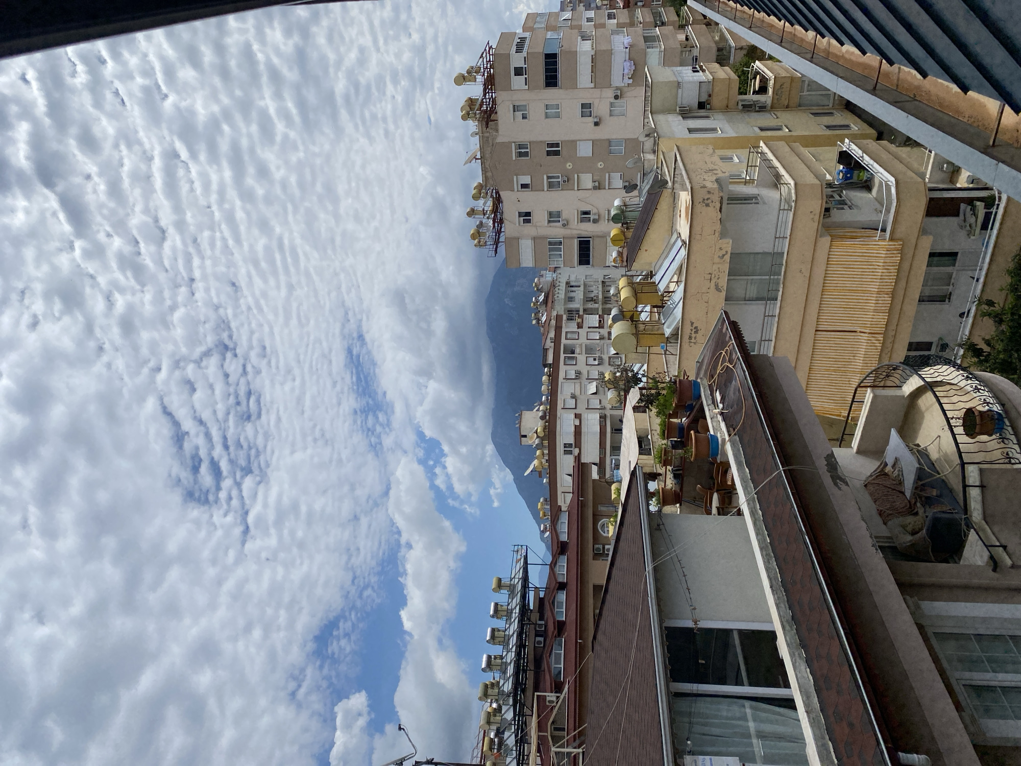 The image shows a view of the city of Alanya, Turkey, from a high vantage point, possibly a balcony or rooftop. The perspective is slightly angled. The sky is partially cloudy with a mix of white and gray clouds. In the distance, a mountain is visible. Several buildings of varying heights are in the foreground and midground, some equipped with solar water heaters or satellite dishes on their roofs. The buildings appear to be residential apartments. There are no people visible in the image. The environment suggests an urban setting, and the architecture is characteristic of a Mediterranean town. The image is rich in details of the cityscape.