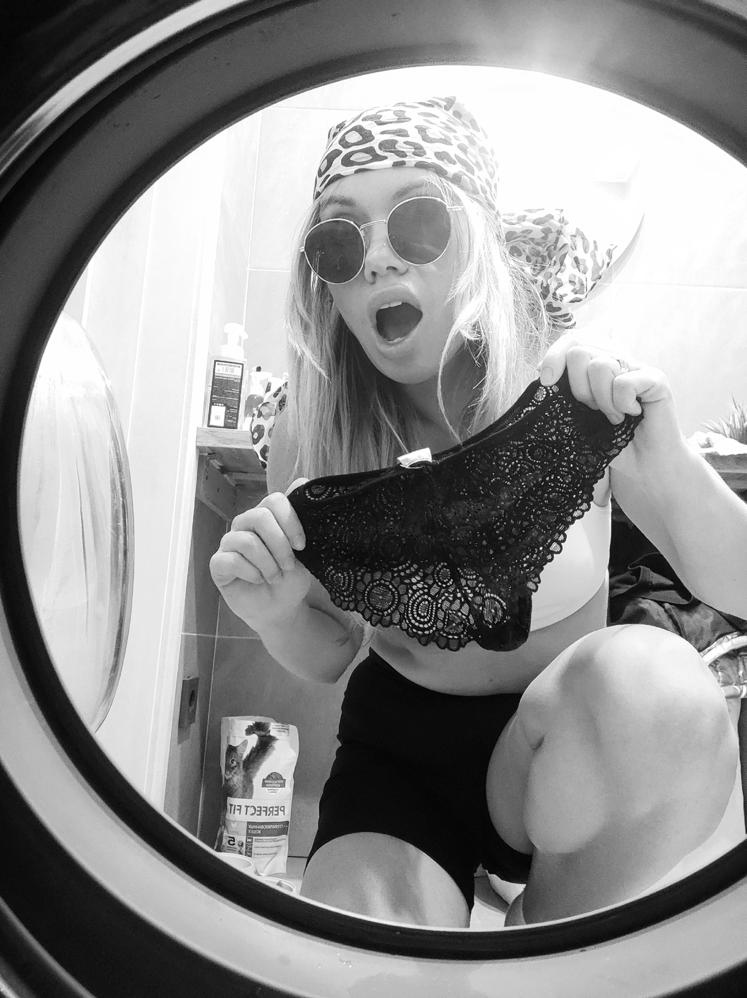 A woman with blond hair, wearing round sunglasses and a leopard-print headscarf, is crouching, with her mouth open in a surprised expression, and holding up a pair of black lace underwear. She is wearing a white top and black shorts. The scene is framed by the circular opening of a washing machine. In the background, there is a tiled wall, a shelf with cleaning products, and a bag of "Perfect Fit" cat food. The location of the image is unknown and cannot be determined to be Votkinsk, Russia.