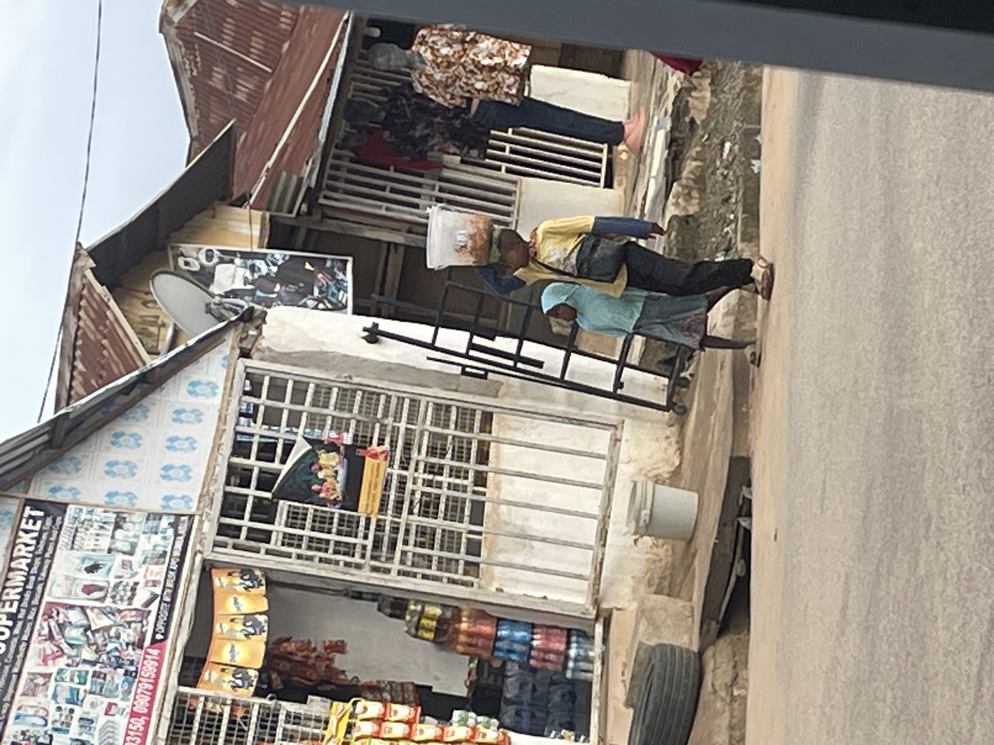 The image presents a street scene in Abuja, Nigeria, specifically near Wuse Abia Market, as indicated by signage. In the foreground, a paved road occupies the lower portion of the frame. On the right, two children walk along a raised, unpaved sidewalk. The taller child, wearing a yellow long-sleeved shirt, dark pants, and sandals, carries a clear plastic bucket filled with food on their head, using their right hand for support, and has a dark cross-body bag. Beside them, a smaller child, dressed in a light blue hooded garment, a patterned skirt, and light sandals, walks to their left.
To the left of the children, on the ground, a black tire rests horizontally, adjacent to a white plastic bucket. A black metal ladder leans against the wall of a building further back. The background consists of several commercial structures. A prominent building on the left features a large sign reading "SUPERMARKET" with phone numbers (0803150, 09079159914) and mentions "OPPOSITE WUSE ABIA MARKET." The upper facade of this building displays blue and white patterned tiles. Its ground-level store entrance is secured by metal grates, revealing stacks of bottled beverages and hanging packaged goods inside. "D-Link" branded banners hang above this entrance. To the right of the supermarket, another structure features a satellite dish and a poster depicting an individual. Further right, a third store entrance showcases two mannequins, one in a floral top and dark pants, the other in a patterned shirt and dark pants. All visible buildings are topped with corrugated metal roofs. The sky above is overcast.
