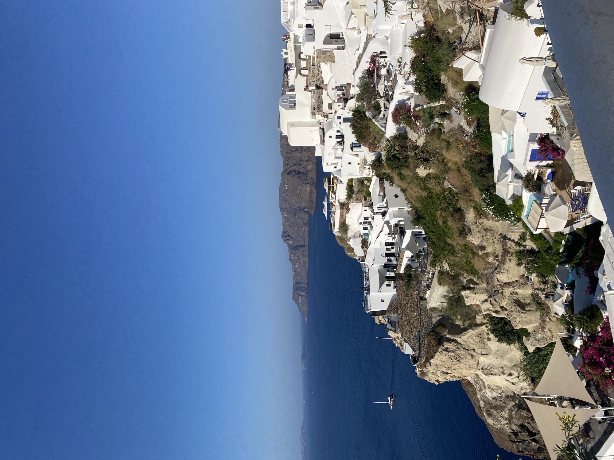 The image depicts a panoramic view of the village of Oia on the island of Santorini, Greece, overlooking the Aegean Sea. The scene is characterized by the iconic white-washed Cycladic architecture, with buildings cascading down a cliffside towards the water. The structures feature domed roofs, narrow passageways, and blue accents on doors and windows, typical of the region.
In the foreground, a white building with a curved roof and blue shutters is visible, adorned with flowering bougainvillea. A dining area with white umbrellas and tables is set up on a lower terrace. Further down the cliff, more white buildings are nestled amongst lush greenery and rocky outcrops.
The deep blue of the Aegean Sea dominates the midground, with a white sailboat sailing in the distance. The coastline of Santorini is visible in the background, characterized by rugged, dark-colored cliffs and smaller islands dotting the horizon under a clear, bright blue sky.
Several people are present, scattered across the rooftops and terraces of the village, suggesting a leisurely atmosphere. The sunlight casts distinct shadows, indicating a bright, daytime setting. The overall impression is one of a picturesque and serene Mediterranean landscape.
