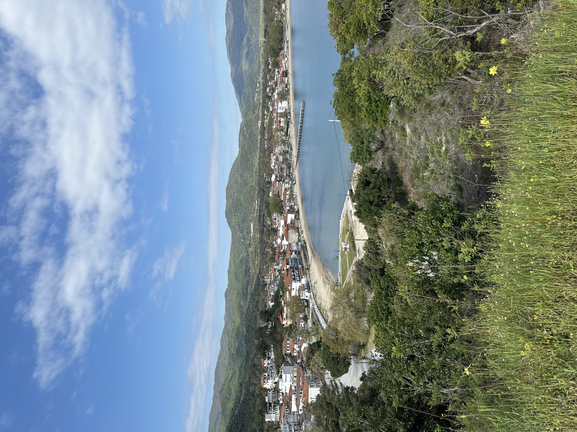 The image captures a coastal cityscape in Ολυμπιάδα, Greece, viewed from an elevated position. The town is nestled along a bay bordered by mountains on one side and the sea on the other. A pier extends into the water. The coastline features a sandy beach alongside buildings and infrastructure. The foreground consists of vegetation, including trees, shrubs, and grassy areas with yellow wildflowers. Above, the sky is blue with scattered white clouds. The image provides a panoramic view of the landscape.