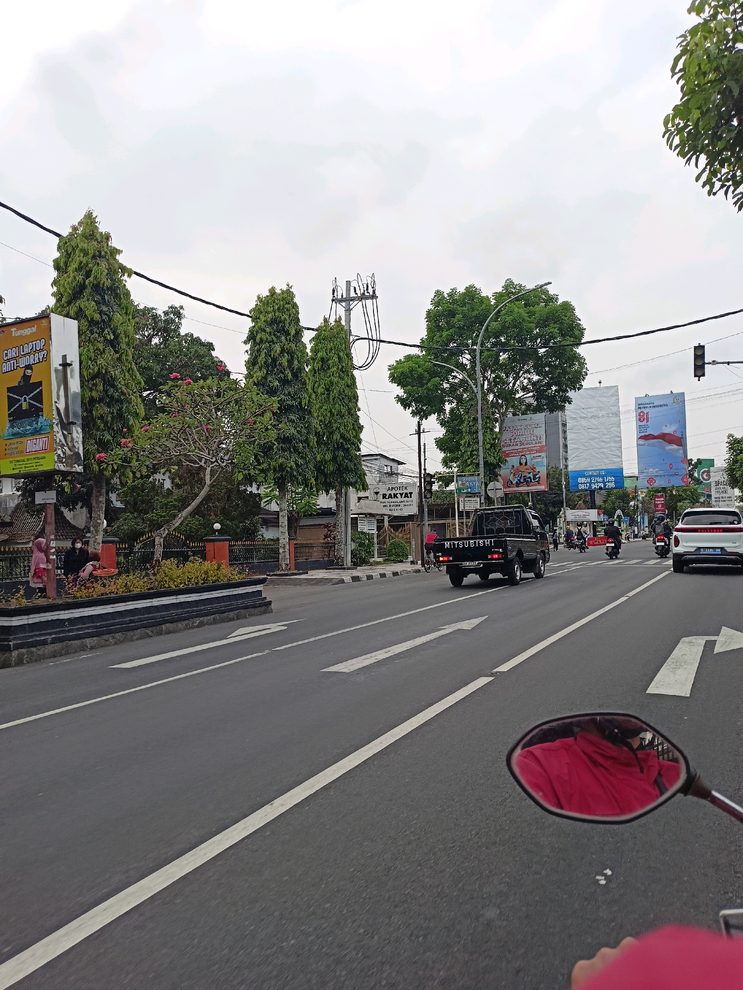 The image captures a street scene in Magelang, Indonesia, on an overcast day. The perspective is from a motorcycle, as evidenced by the rearview mirror reflecting a person wearing a pink garment. The road is asphalt with white lane markings. On the left side of the road, a billboard advertises "Tunggal Cari Laptop Anti-Worry." Several tall, slender trees line the median, with a larger, leafy tree further back. A black Mitsubishi pickup truck is in the foreground, heading away from the camera. Behind it, several motorcycles and a white car are also on the road. In the distance, more billboards and buildings are visible, including a traffic light. The overall atmosphere is that of a typical urban street in Indonesia.