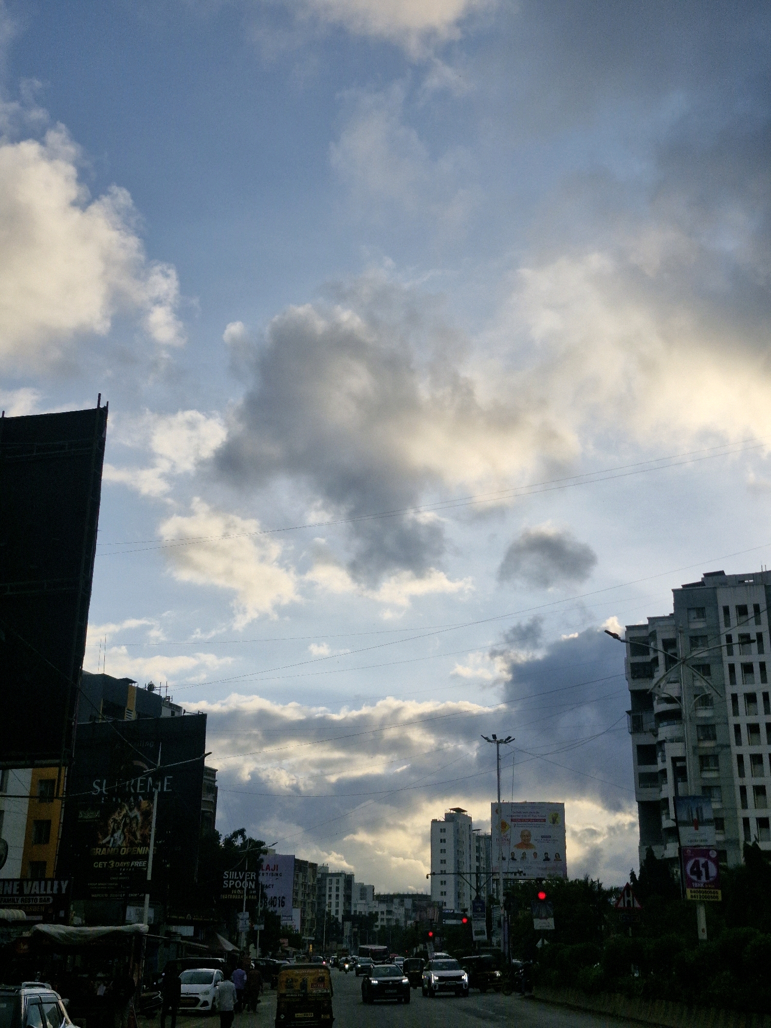 The image depicts a street scene in Tathawade, India, under a cloudy sky. Tall buildings, including a dark, unfinished structure on the left and a modern apartment building on the right, dominate the background. Several billboards are visible, advertising various businesses and events, such as "Supreme" and "Silver Spoon." The road is lined with trees and traffic lights. Multiple vehicles, including cars, an auto-rickshaw, and what appears to be a motorcycle, are present on the road, suggesting moderate traffic. A few pedestrians are visible on the sidewalk, near a market stall. The sky displays a mix of light and dark clouds, with the sun partially visible through the overcast conditions, casting a subdued light on the scene.
