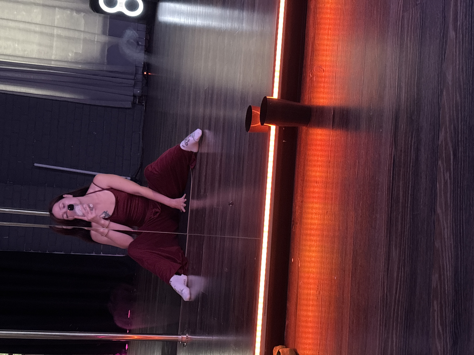 The image shows a person sitting on a wooden floor in what appears to be a dance studio. The person is wearing a maroon jumpsuit and white sneakers and is holding a phone to take a selfie in a mirror. The studio has a large mirror reflecting the person and the studio environment. There are two dark cups on the floor in front of the mirror, near a strip of orange LED lighting. The background includes dark curtains and a brick wall. The overall lighting is dim, with the orange LED strip providing a prominent light source. There is no visible indication of the location being in Naberezhnye Chelny, Russia.