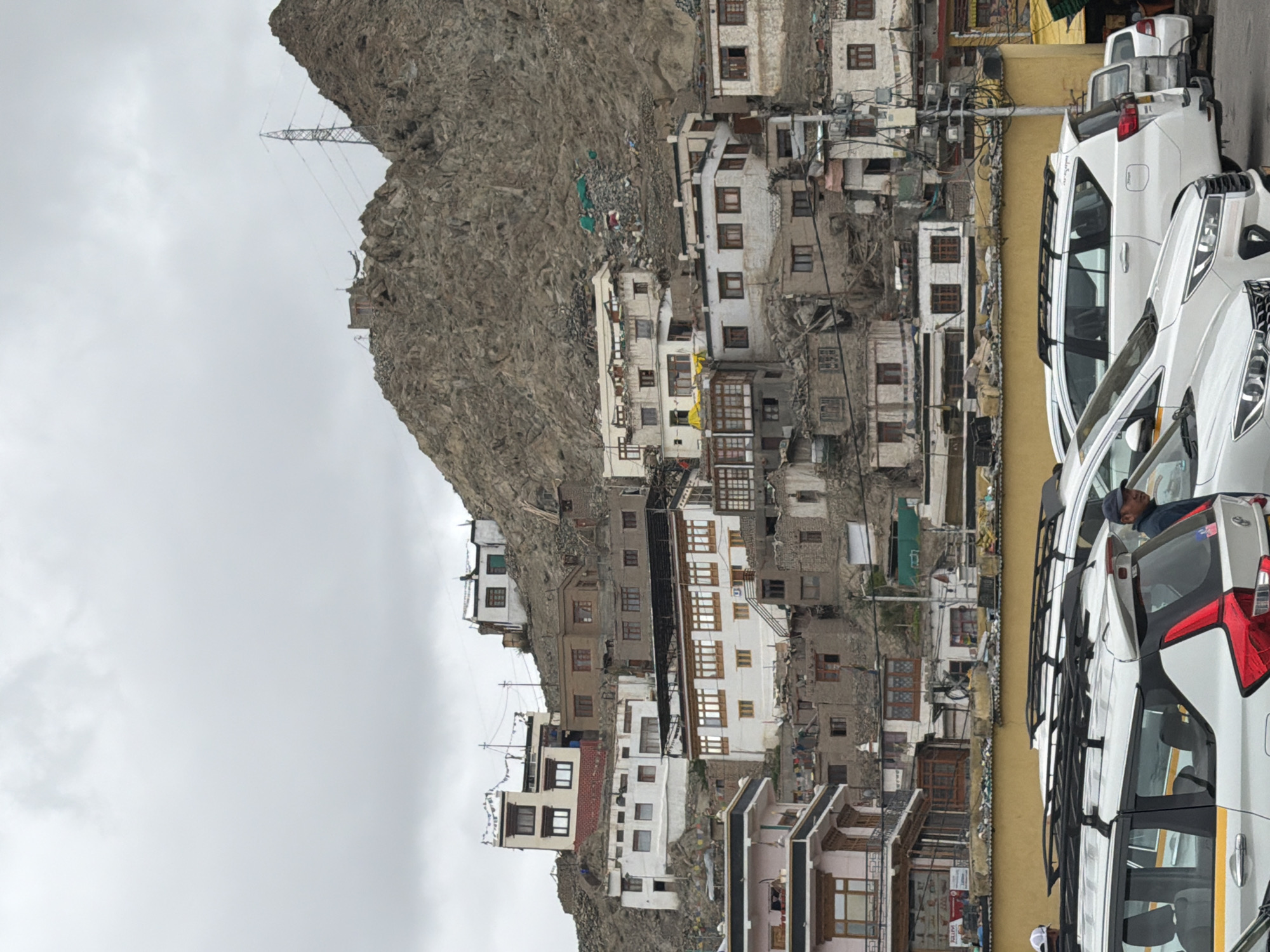 This image captures a view of the city of Leh, India, a mountainous region known for its Tibetan Buddhist culture and monasteries. The scene is dominated by a densely packed hillside settlement, with buildings of various sizes and styles clinging to the rocky terrain. The architecture appears to be a mix of traditional and modern, with whitewashed walls and wooden accents evident on many structures. Some buildings feature prayer flags, adding a spiritual element to the urban landscape.
In the foreground, several white vehicles, likely taxis or tourist vans, are parked in a lot. The vehicles have roof racks, suggesting they are used for transportation in the rugged region. A man wearing a dark baseball cap stands near the rear of one of the white vehicles, looking towards the right of the frame. The sky is overcast with heavy clouds, casting a diffused light over the scene. In the background, a large, rocky mountain rises behind the settlement, with a power line tower visible on its peak. The overall impression is of a vibrant, yet rugged, community nestled within a dramatic natural environment.