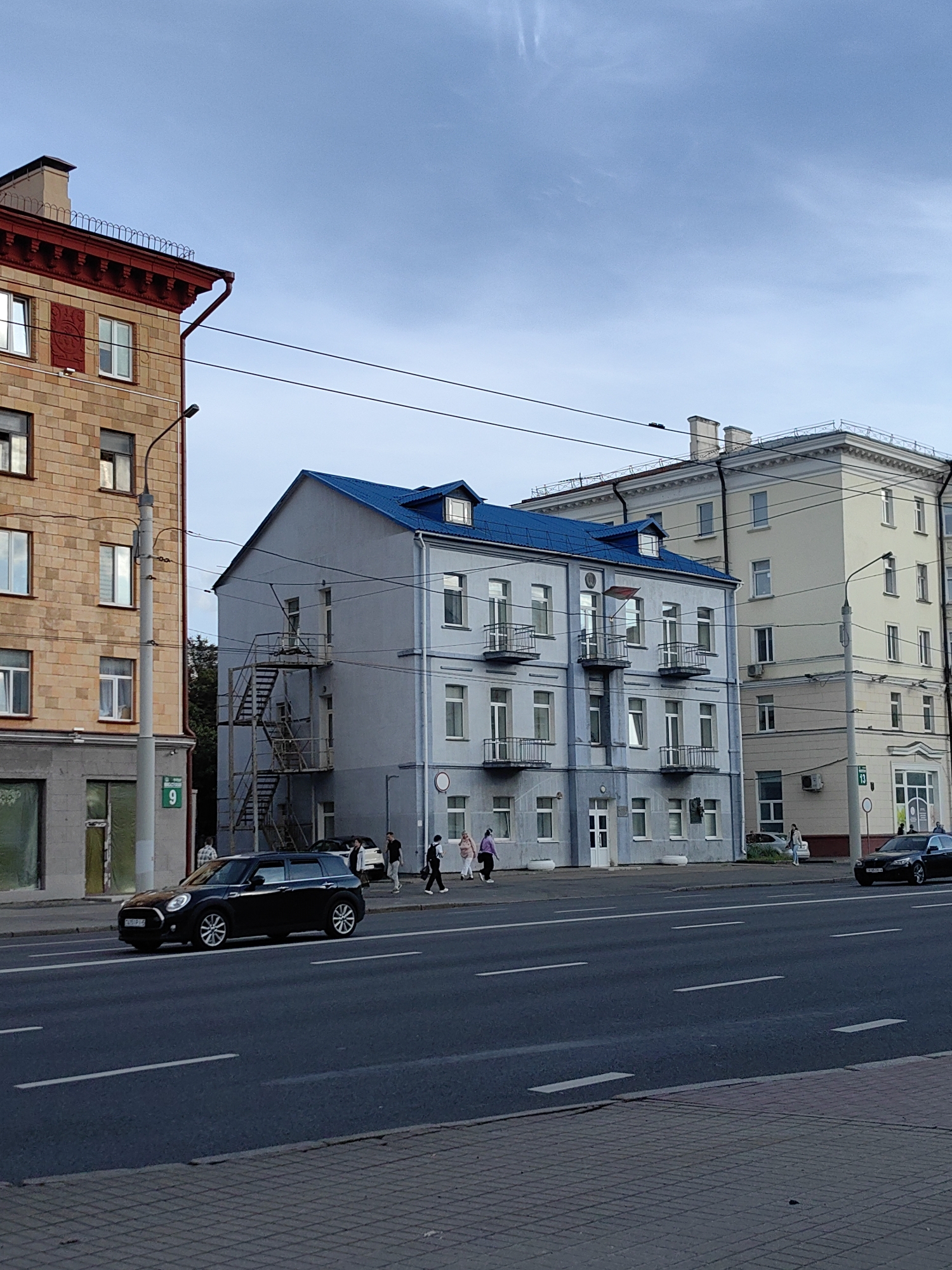 The image depicts a street scene in Minsk, Belarus, likely during the daytime under a cloudy sky. On the left, a multi-story building with a light brown brick facade and a red decorative cornice is visible. To its right, a narrower, light blue building with a blue roof and external fire escapes is situated. Further to the right, a taller, cream-colored building with a classical facade is present. A paved road with lane markings runs across the foreground, with a sidewalk and cobblestone pattern bordering it. Several people are walking on the sidewalk, and two cars are visible on the road. One is a dark-colored Mini Cooper hatchback in the left lane, and another is a dark sedan in the right lane. Streetlights and power lines are present throughout the scene, along with street signs. The environment appears to be urban, with architecture suggesting an older part of the city.
