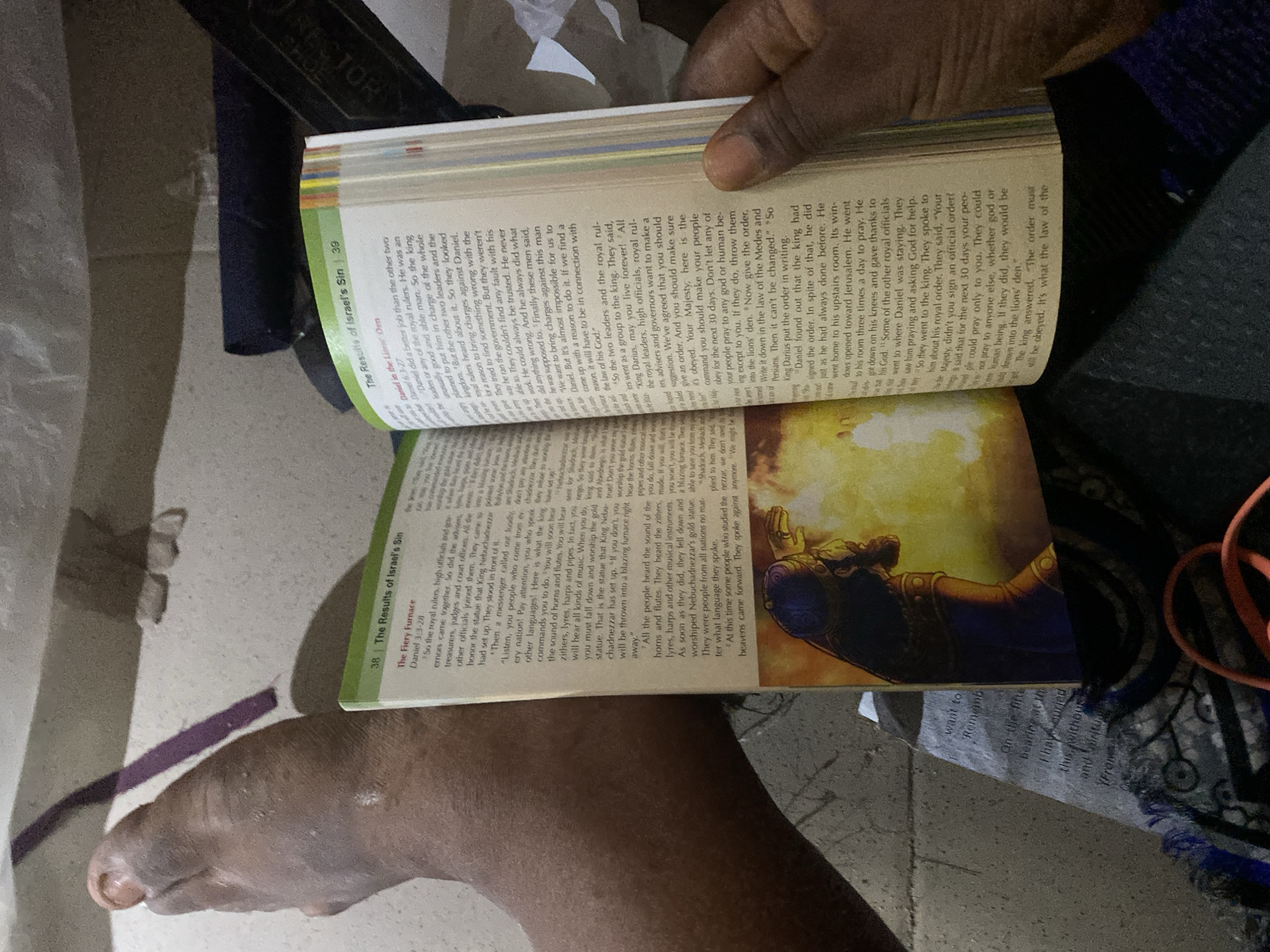 In Jauro Yinu, Nigeria, an image captures a person's foot and hand resting on an open book. The book displays text and an illustration, suggesting a religious or educational context. The surrounding environment is dimly lit, with the focus on the book and the individual's limbs. The overall impression is one of quiet study or reflection in a personal space.