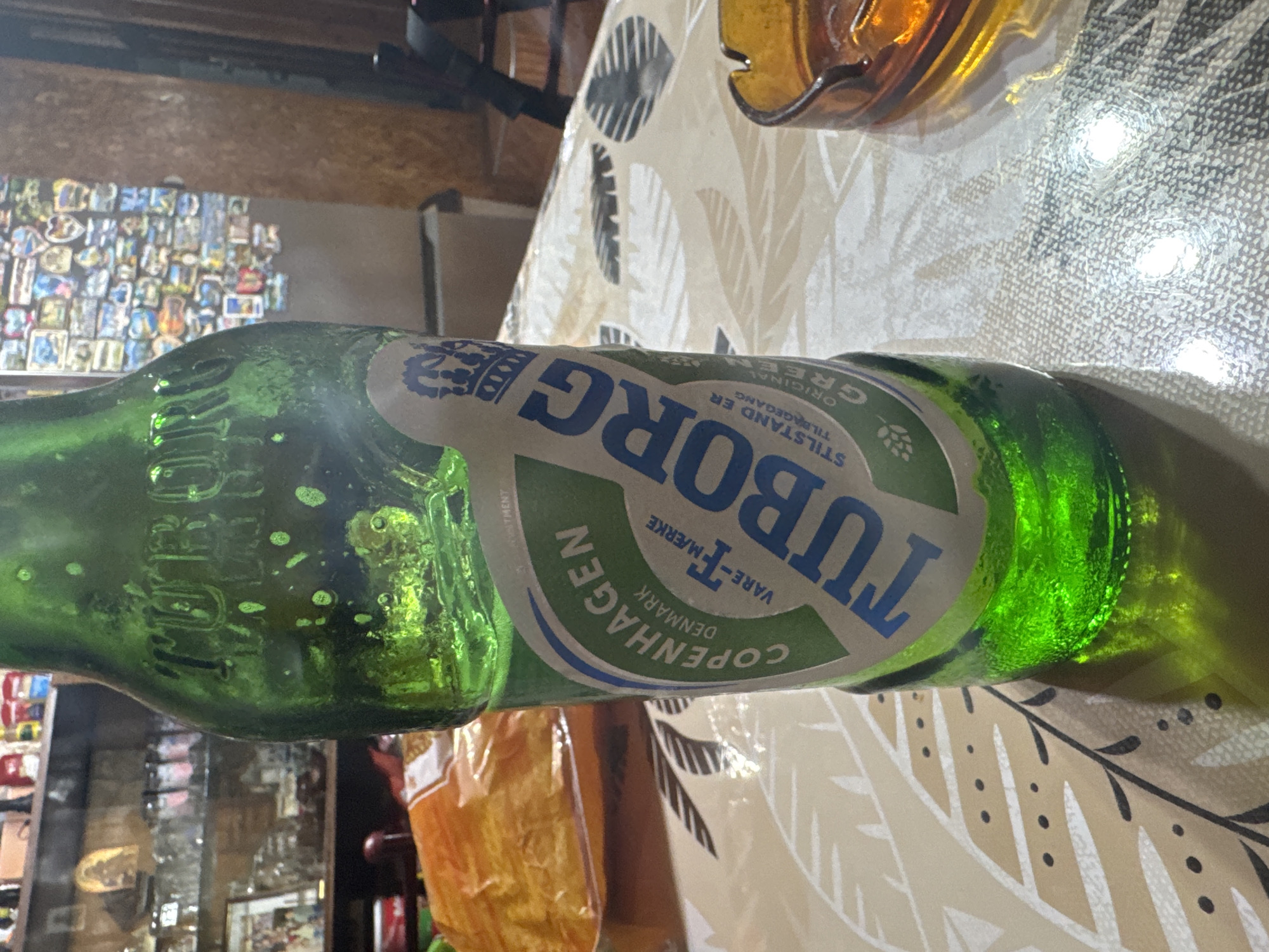 The image displays a green glass bottle of Tuborg beer resting horizontally on a table. The bottle's label features the prominent blue text "TUBORG," along with "COPENHAGEN," "DENMARK," "VARE-T-MÆRKE," "ORIGINAL," "GREEN," and the Danish phrase "STILLSTAND ER TILBAGEGANG" (meaning "Standing still is going backwards"), accompanied by a hop cone illustration.
The table is covered by a light-colored tablecloth patterned with dark grey or black leaves and dots. Partially visible in the upper right is an amber-colored glass object, possibly an ashtray. The background is mostly out of focus but shows a wall adorned with a dense collage of numerous small pictures or magnets. Other blurry elements include a brown wooden surface in the far upper left and an orange or yellow object, possibly packaging, in the lower left.
The scene depicts an indoor setting, likely a casual establishment or home in Sarnevo, Bulgaria, under artificial lighting. No people are visible, and no specific activity is taking place, presenting a still life composition.