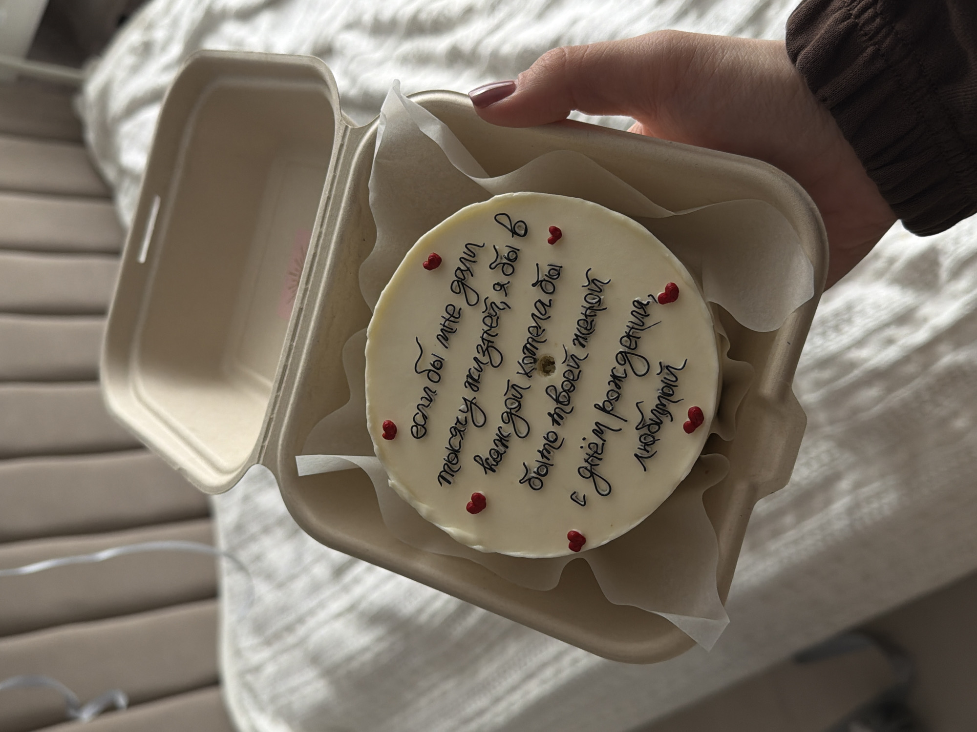 A close-up, high-angle shot captures a hand holding an open, beige, disposable takeout cake box containing a small, round white cake. The cake is adorned with several small, red heart-shaped sprinkles. The background is softly blurred, showing a light-colored, possibly knitted or textured fabric, suggesting an indoor setting like a bed or blanket.
The hand, with fingernails painted a reddish-brown, holds the box carefully. Part of an arm wearing a brown, ribbed sleeve is also visible. The cake is the central focus, and its white surface features handwritten text in black, along with the heart sprinkles.
The text on the cake, written in Cyrillic, translates to a romantic birthday message: "If I were given a thousand lives, I would want in each of them to be your wife. Happy Birthday, my love." This indicates a celebration, specifically a birthday, and a strong affectionate or romantic relationship between the giver and the recipient of the cake. The overall mood is one of love and celebration. The scene takes place indoors, likely in Reshetnikovo, Russia, as per the provided context.