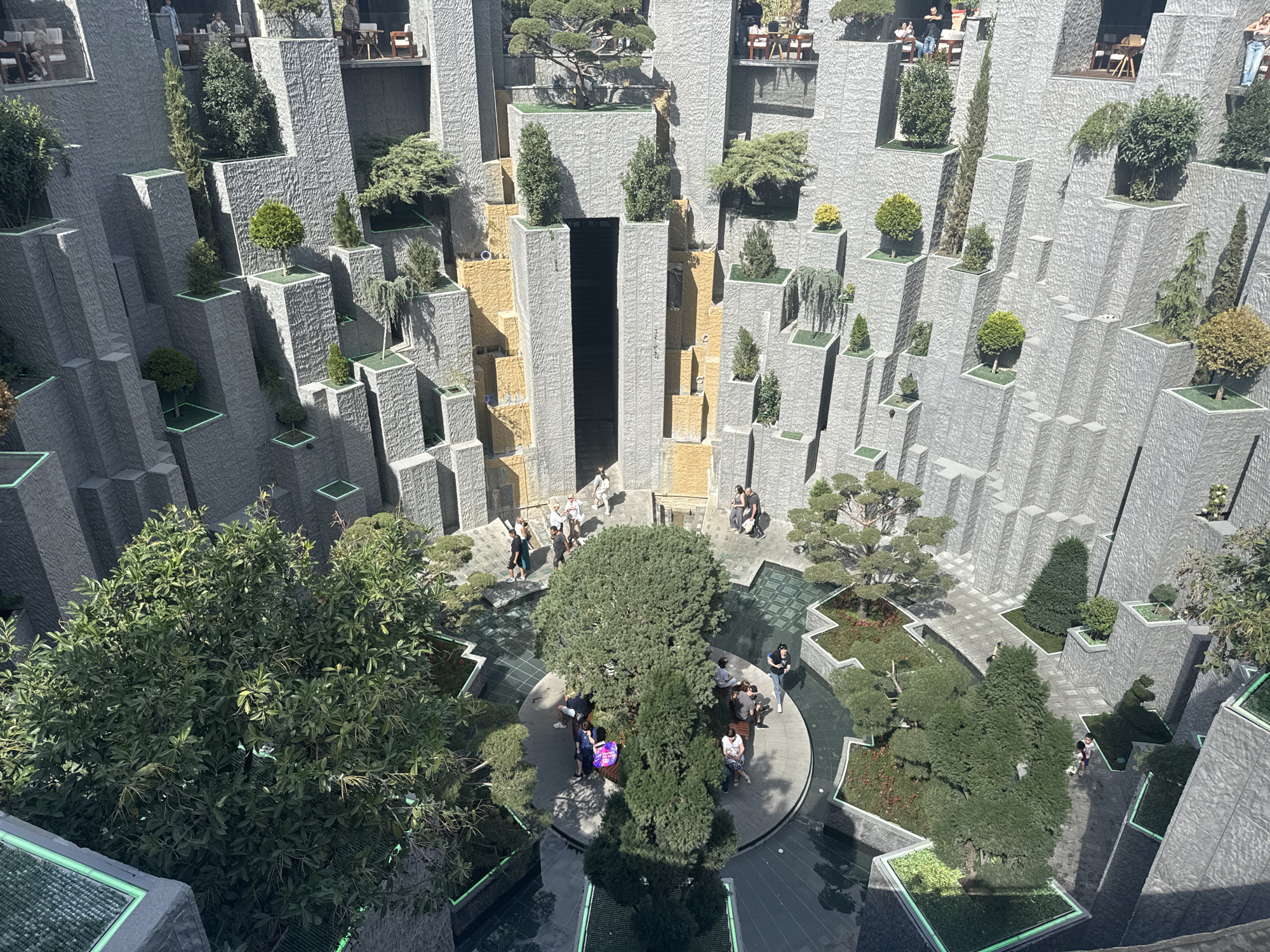 The image depicts a multi-level outdoor courtyard, possibly a public space or part of a building complex, featuring a striking architectural design with numerous angular, stacked stone elements. These elements are arranged to create vertical planting spaces for various trees and shrubs, forming a tiered, garden-like environment. In the lower central area, several people are visible, some seated on benches around a circular garden bed with a large tree. Other individuals are walking along pathways and in groups. The overall aesthetic is modern and geometric, with the grey stone contrasting with the vibrant green of the vegetation. This scene is identified as being located in Krasnodar, Russia.