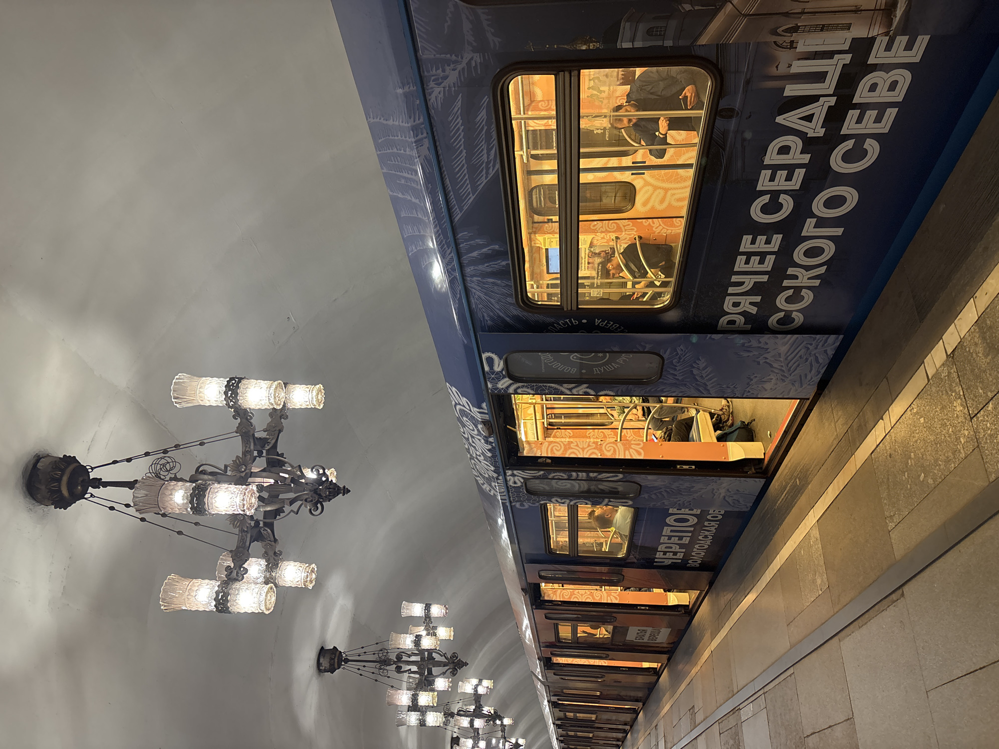 The image captures the interior of a Moscow Metro station in Russia. A blue metro train decorated with white designs and Cyrillic text is stopped at the platform. People are seated inside the train, visible through the windows. Overhead, elaborate chandeliers illuminate the station. The platform is made of stone tiles. The station has a curved, white ceiling.