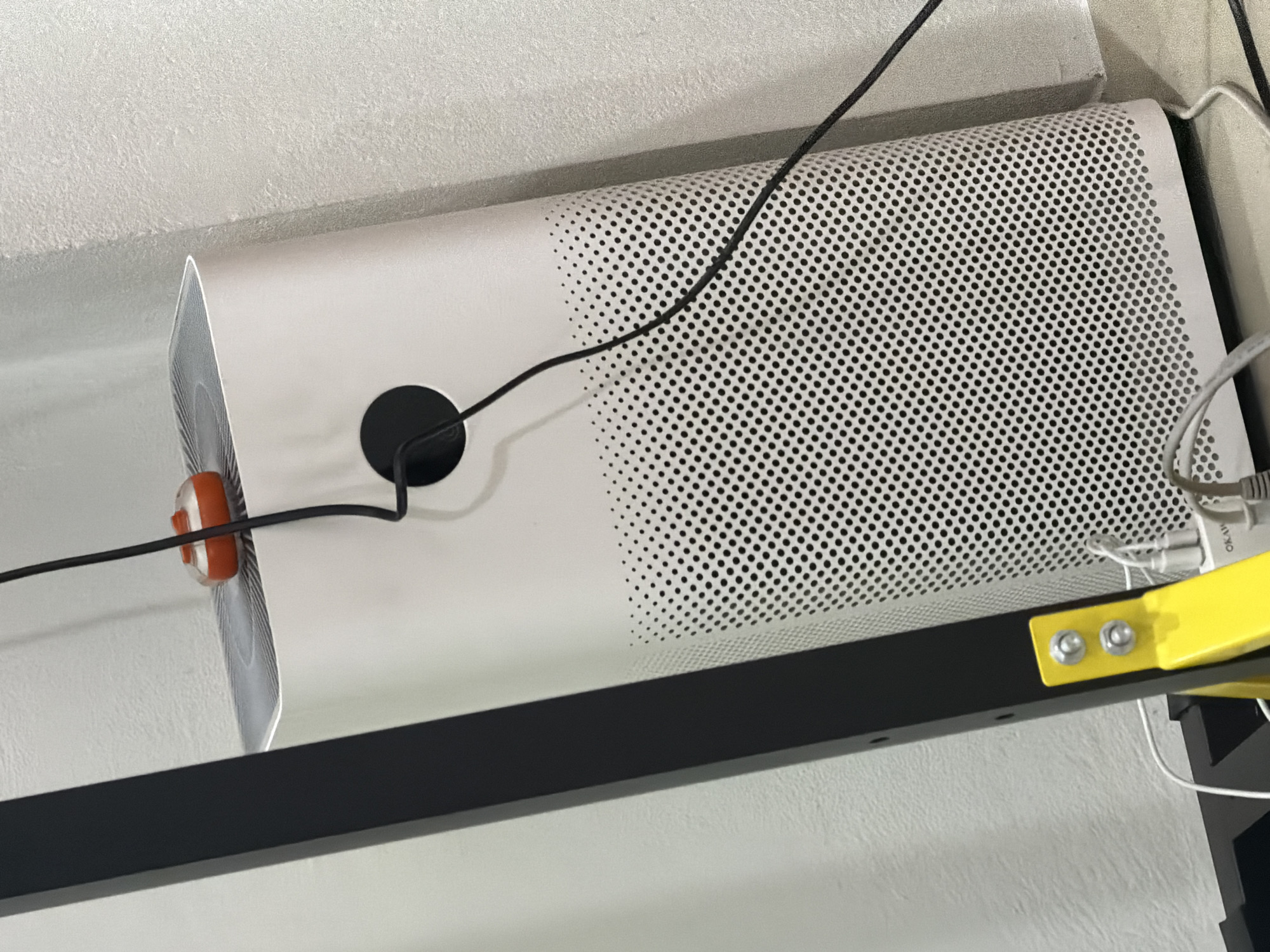 An indoor scene, likely in Baan Mae Nam, Thailand, captures a white air purifier situated next to a black metal stand. The air purifier has a perforated grill pattern on its front and top, and a black circular vent on its upper front panel. A black power cord runs from the top of the air purifier, where a small orange and clear object, resembling a curling stone, is placed on top of the unit. The scene is illuminated by ambient light, casting soft shadows. The background consists of a textured white wall. To the left of the air purifier, a yellow metal bracket with two silver bolts is visible, attached to the black stand. Various white and grey electrical cables are present near the base of the air purifier and the yellow bracket.