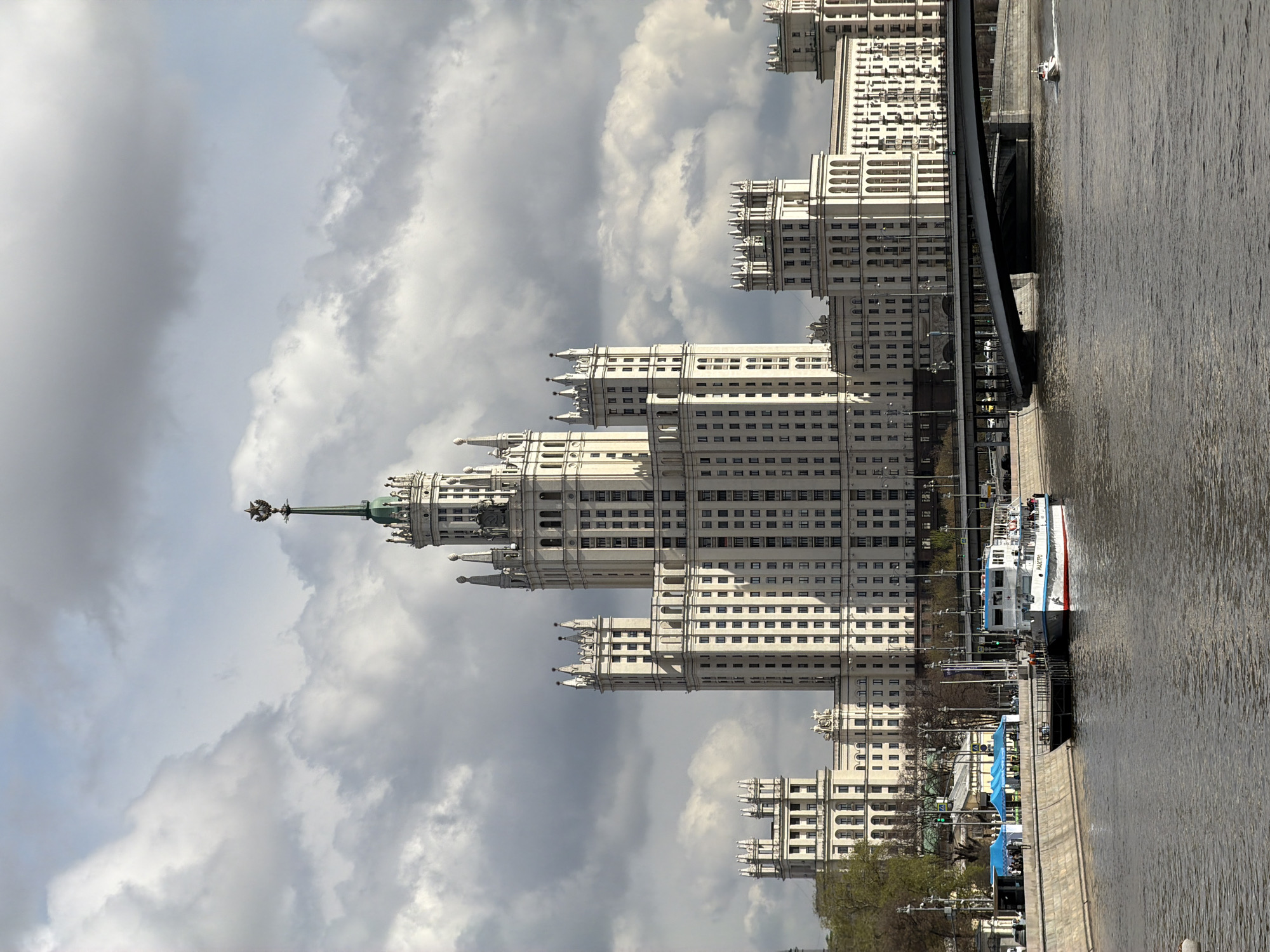 This image, when correctly oriented, depicts a vibrant urban scene along a wide river in Moscow, Russia. The most prominent feature is a large, ornate, light-colored skyscraper in the Stalinist Empire style, likely one of Moscow's "Seven Sisters," dominating the left side of the frame. This majestic building features multiple tiers of architectural elements, numerous windows, and a distinctive green spire topped with a star-like ornament.
In the foreground and stretching to the right, the Moskva River flows. A white and blue passenger boat, clearly labeled "МАЭСТРО" (MAESTRO), is docked at a pier on the riverbank. Further along the river, a smaller white boat is seen moving through the water. A dark bridge spans the river, partially visible between the main building and the water. The sky is largely overcast with impressive grey and white clouds, indicating a cloudy daytime setting. Some trees with early spring foliage line the riverbank, adding a touch of green to the concrete structures. The overall impression is one of a grand city under a dramatic sky.