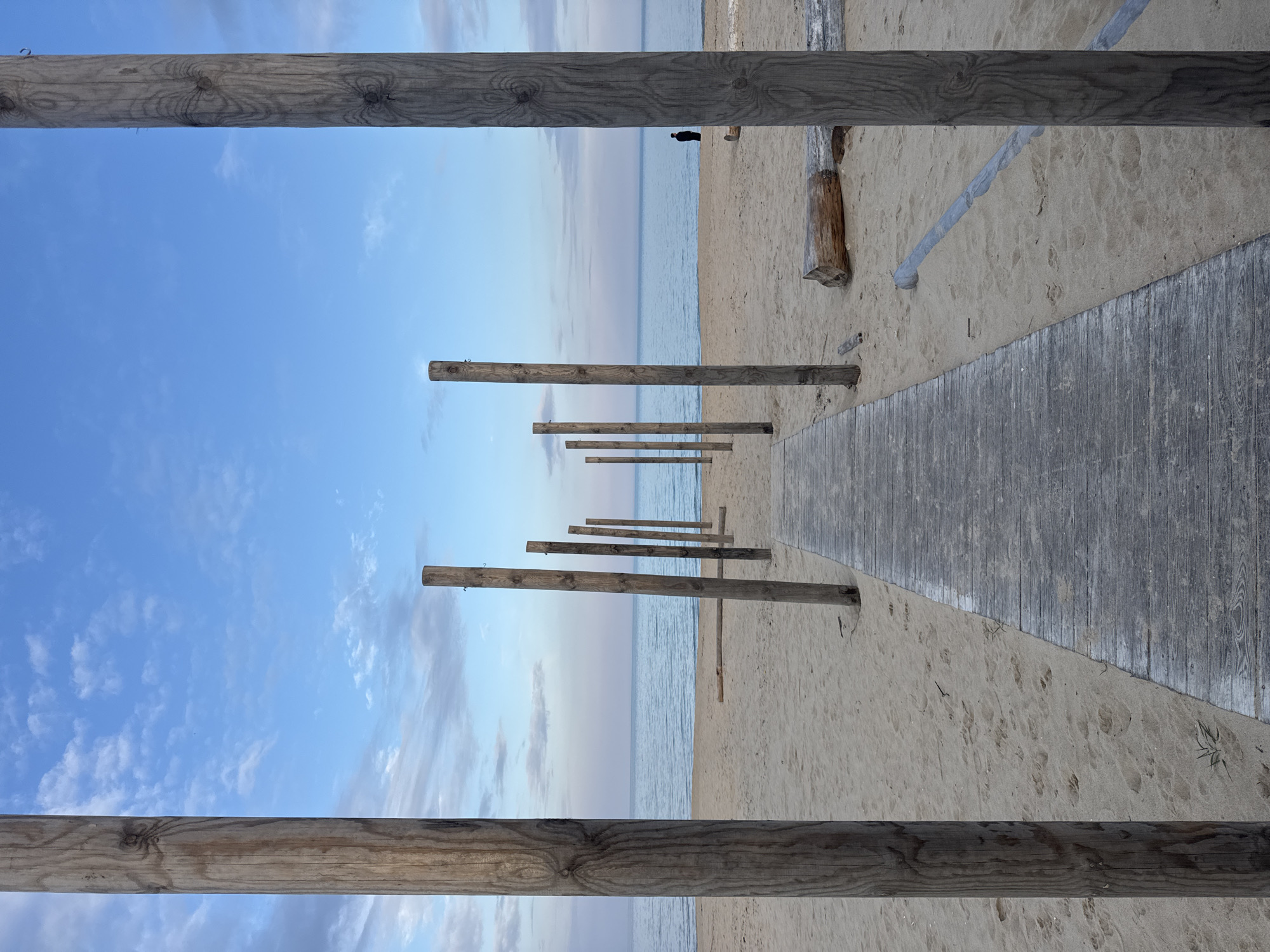 The image shows a beach scene, likely in Kranevo, Bulgaria. The composition is framed by wooden posts and beams, creating a pathway effect. A wooden boardwalk stretches diagonally across the right side of the frame, leading towards the sea. The beach sand is light-colored. Several wooden posts stand perpendicular to the boardwalk, creating a visual barrier or marker along the beach. The sea is visible in the background, with a person standing near the water's edge. The sky is blue with scattered clouds. A piece of driftwood is present on the beach.