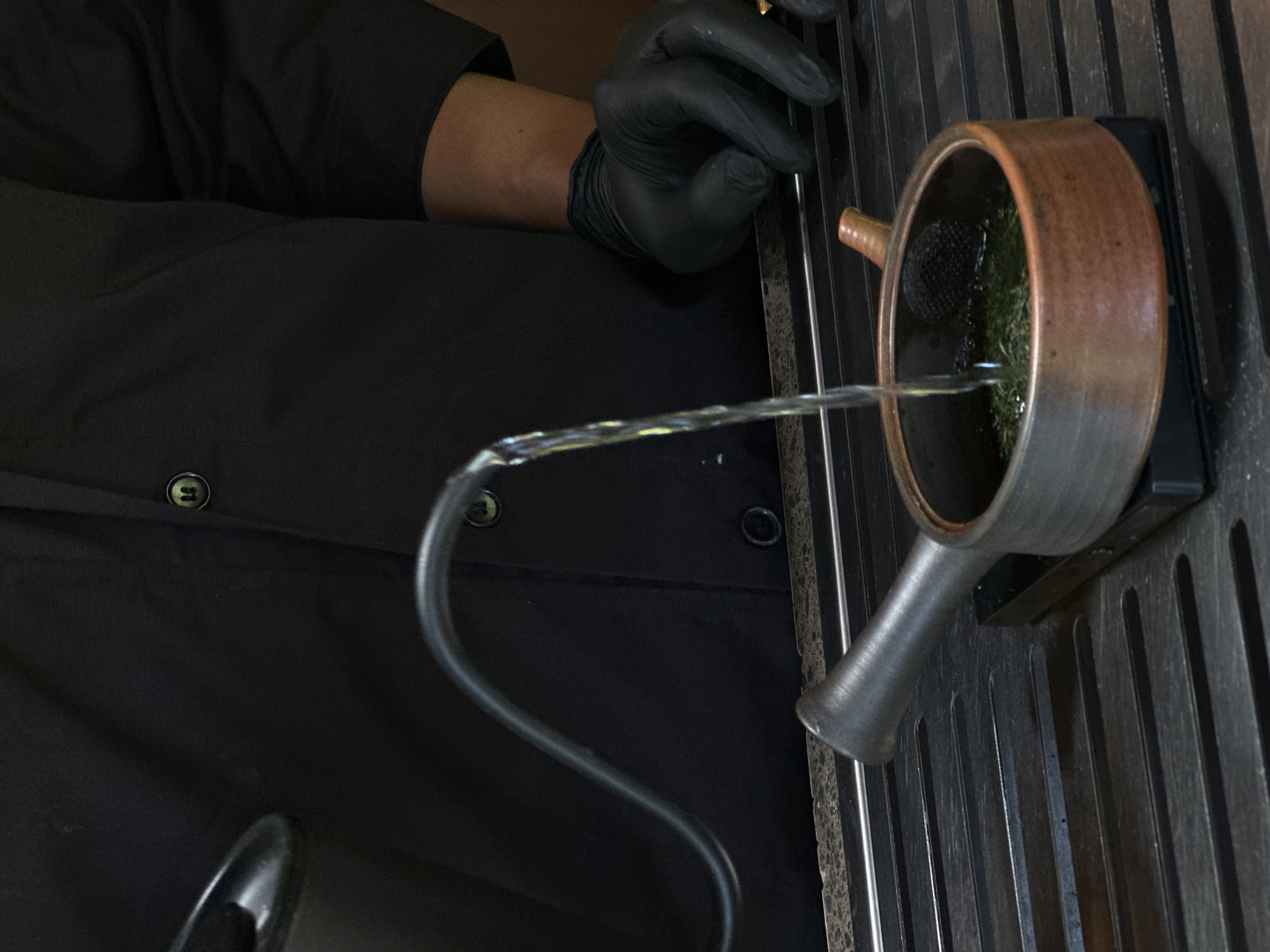 A close-up shot shows a gloved hand holding a flexible metal tube that is pouring water into a ceramic pot filled with green leafy material and a black mesh. The pot is resting on a dark, slotted metal surface. To the left, a person in a black button-down shirt is visible. The origin of the photo is traced to Ubud, Indonesia.
