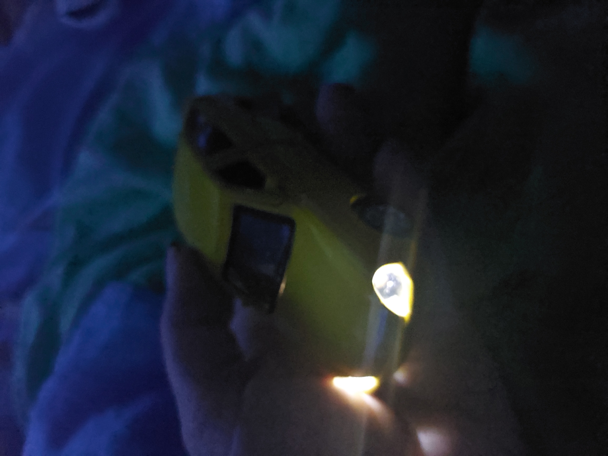 The image presents a close-up, low-light scene featuring a person's hand holding a bright yellow, light-emitting object. The object appears to be a small device, possibly a toy car or a portable gadget, and is equipped with two distinct light sources: a prominent, bright light at its front, resembling a headlight, and a smaller, yellowish light positioned beneath or towards the rear. A dark, rectangular panel or screen is also visible on the side of the yellow object.
The background is dark and out of focus, indicating that the photograph was taken in a dimly lit environment, likely at night or in a dark room. On the left side of the frame, soft textures of fabric in shades of blue and teal/green can be discerned, suggesting a bedspread, blanket, or other soft furnishings. The hand holding the object is partially illuminated by the device's glow, casting a warm light.
The central activity is the holding and operation of this illuminated object. No specific event or complex action is depicted beyond this interaction with the light-emitting device. There are no discernible text elements on the object or within the scene. While the location context is provided as Chaikovsky, Russia, the visual content of the image does not offer specific cues to identify this geographic location. The overall impression is one of quiet focus on the illuminated device in a dark, intimate setting.