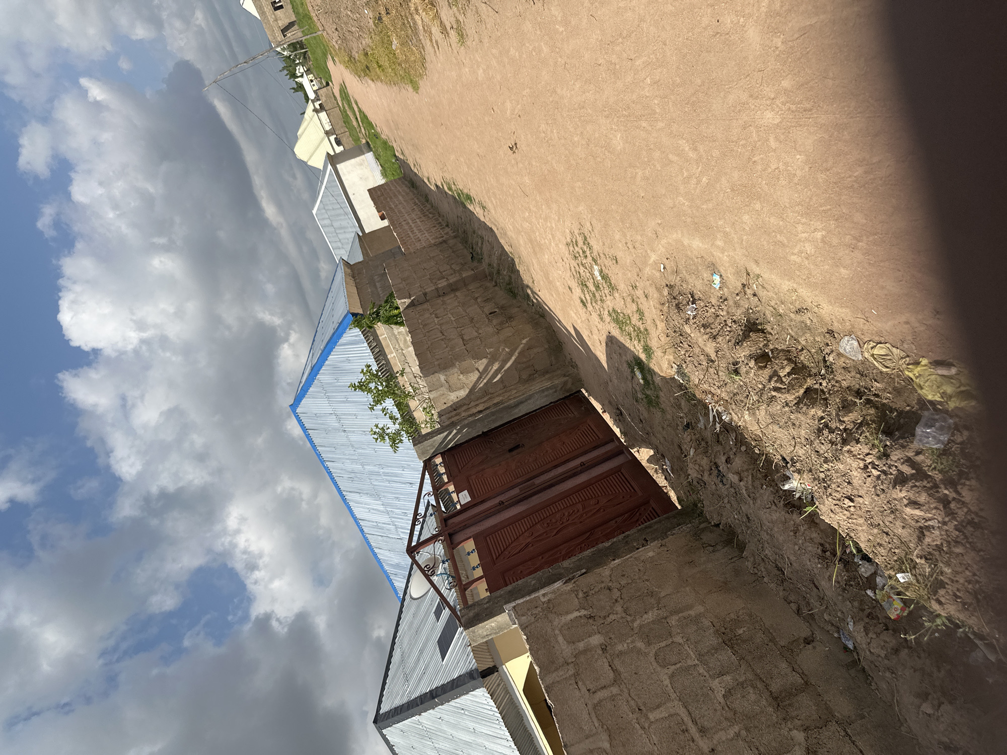 The image depicts a street scene in Jalingo, Nigeria, characterized by a dirt road leading into a residential area. The buildings in the background have corrugated metal roofs, with some featuring blue trim. A prominent feature in the foreground is a red metal gate, partially open, set within a brick wall. The ground along the road and adjacent to the wall is unpaved and shows signs of some scattered debris. The sky above is partly cloudy.