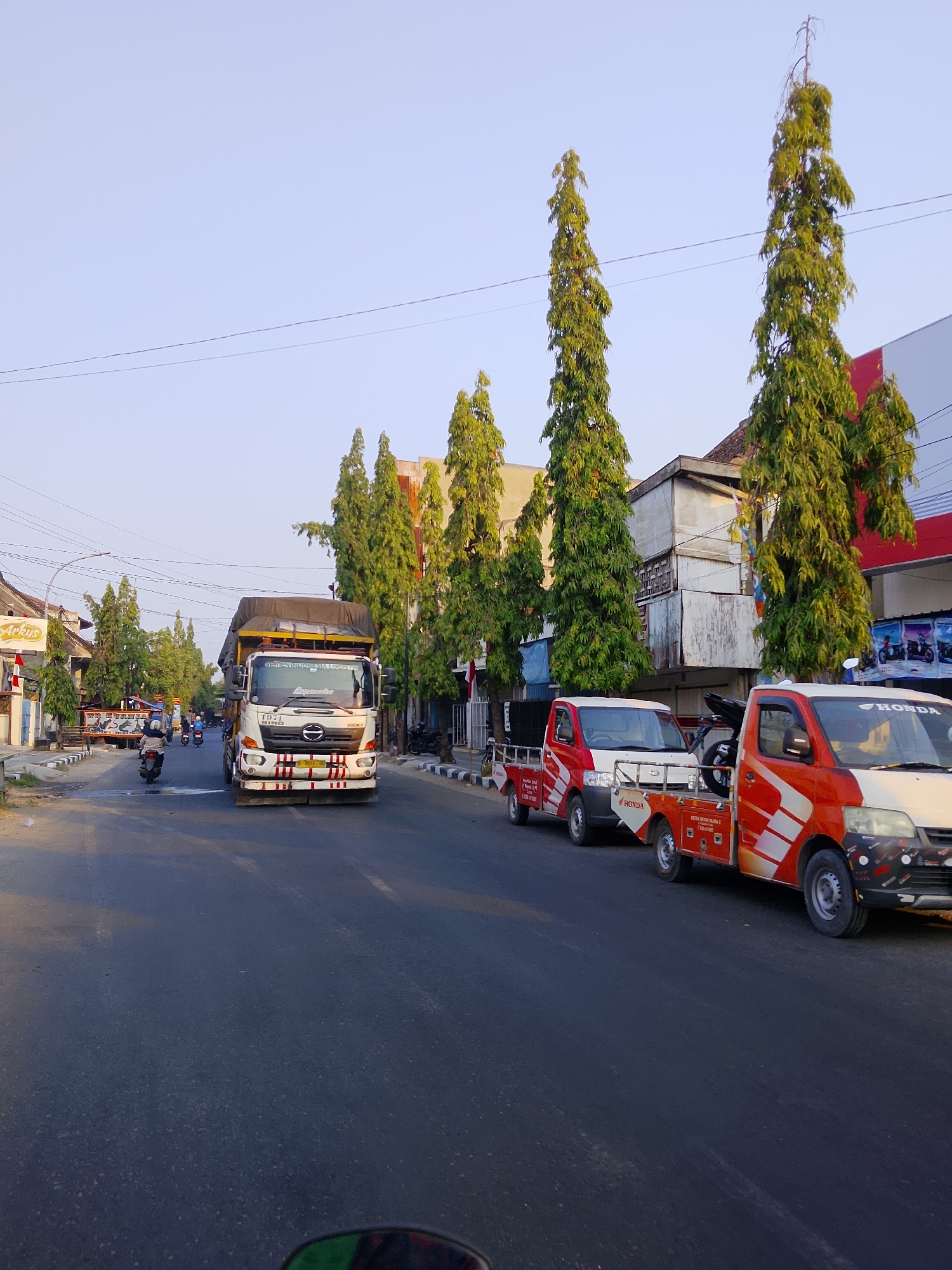The image depicts a street scene in Sambong, Indonesia, with a clear sky overhead. On the right side of the street, two red and white utility vehicles are parked. The vehicle in the foreground has "HONDA" branding visible on its side. Further down the street, a large white truck with a yellow tarp-covered cargo bed is in motion. Several tall, slender trees with green foliage line the street. On the left side, a few motorcycles are visible, one of which is being ridden. Buildings with various architectural styles, some appearing commercial with visible signage, flank the street. The road surface is asphalt with visible lane markings. The overall atmosphere suggests a typical day in a bustling town.